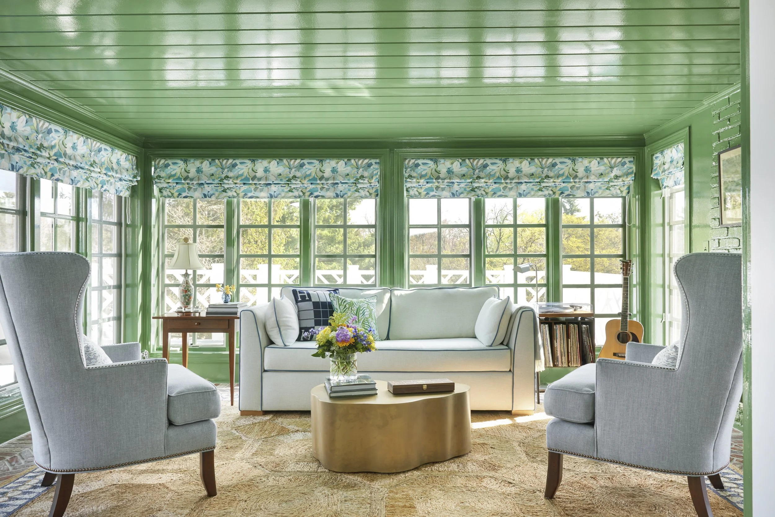 Sunroom with green walls and ceiling, white sofa, two gray armchairs, floral curtains, coffee table with flowers, guitar, and vinyl record collection.