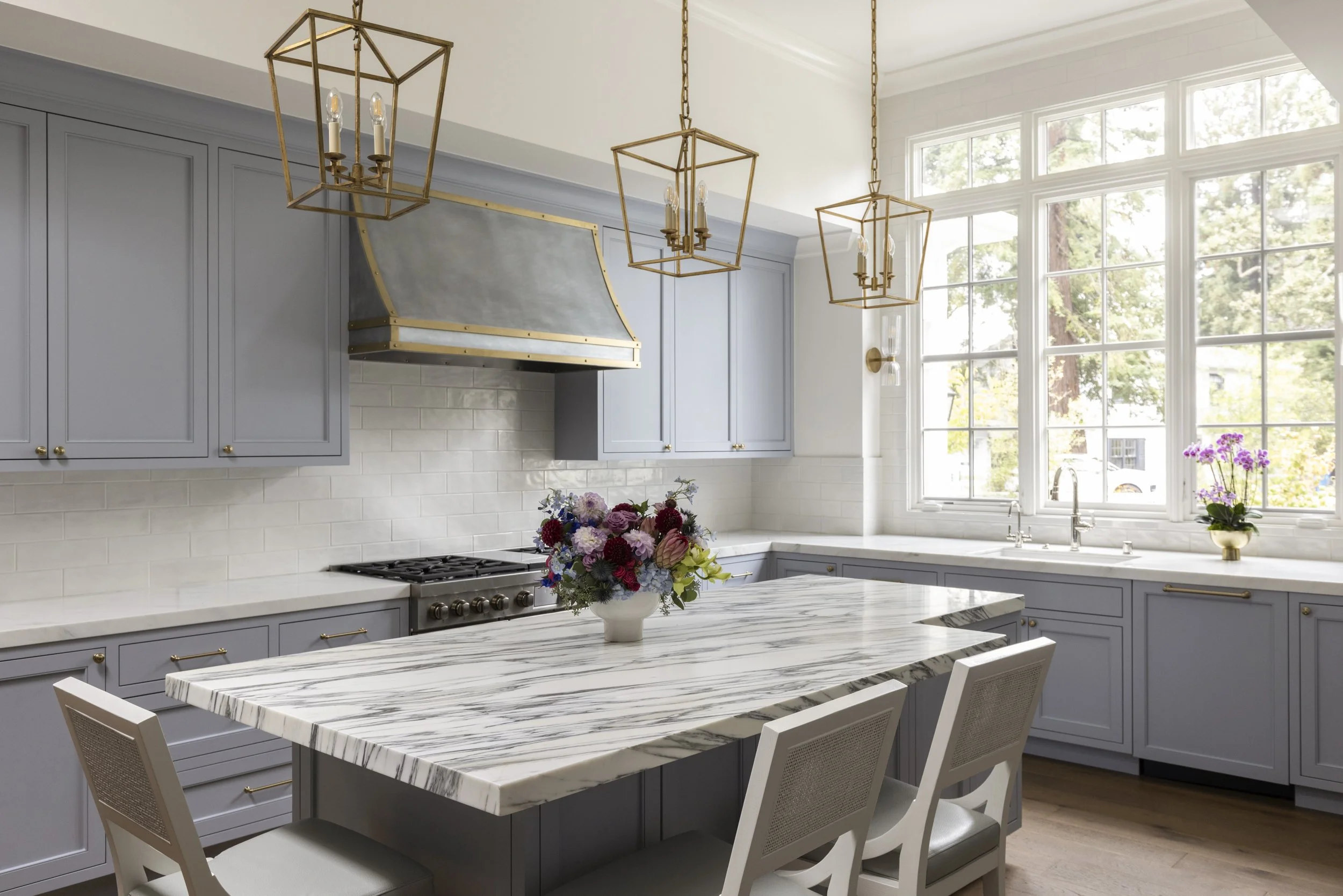 Bright kitchen with light blue cabinets, a marble island, and large windows with a view of trees outside. There is a flower vase on the island and another on the windowsill, with three hanging pendant lights above.