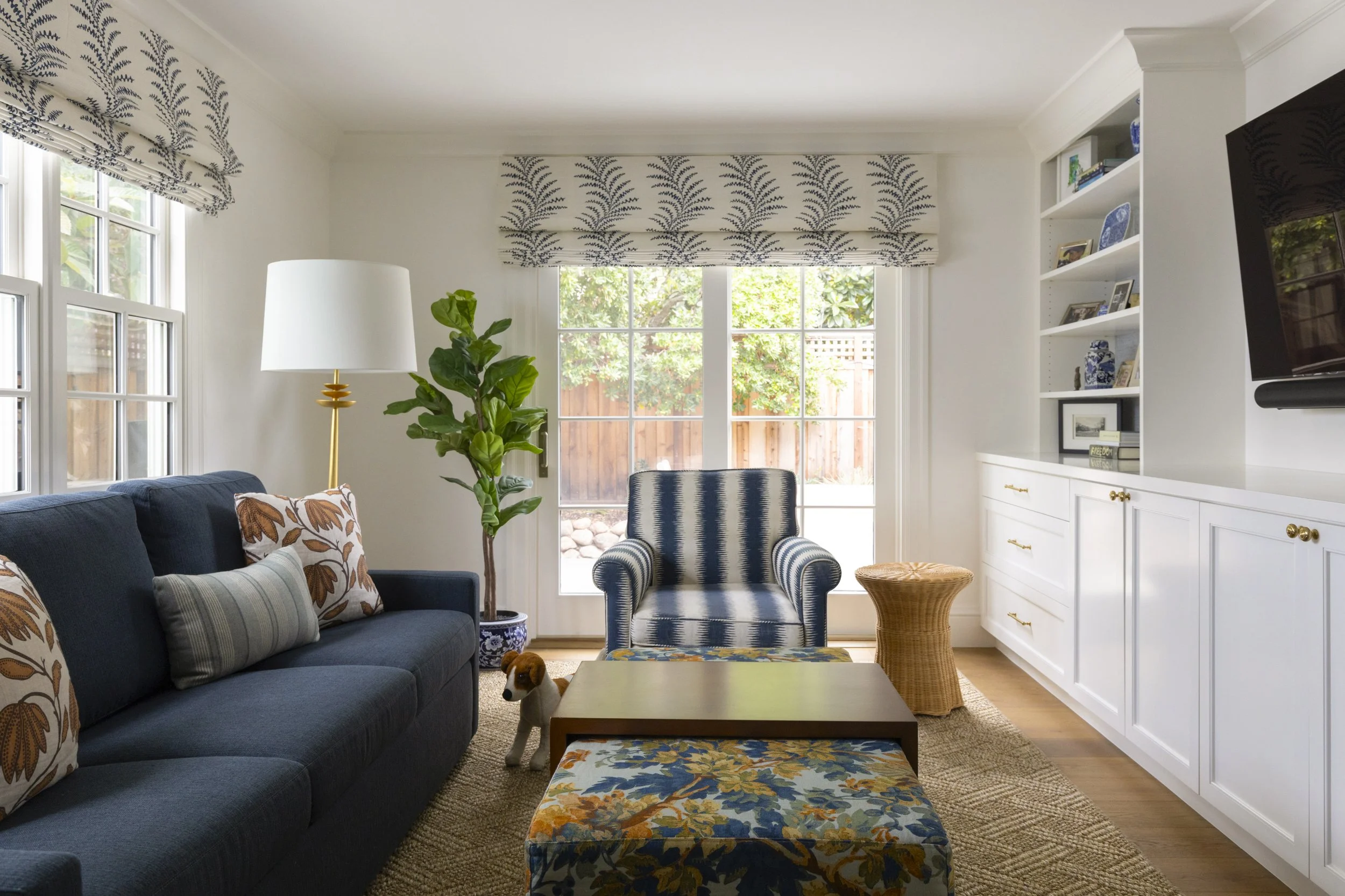 Living room with navy sofa, patterned throw pillows, striped armchair, glass door, white built-in shelves, plants, wooden coffee table, wicker side table, flat-screen TV.