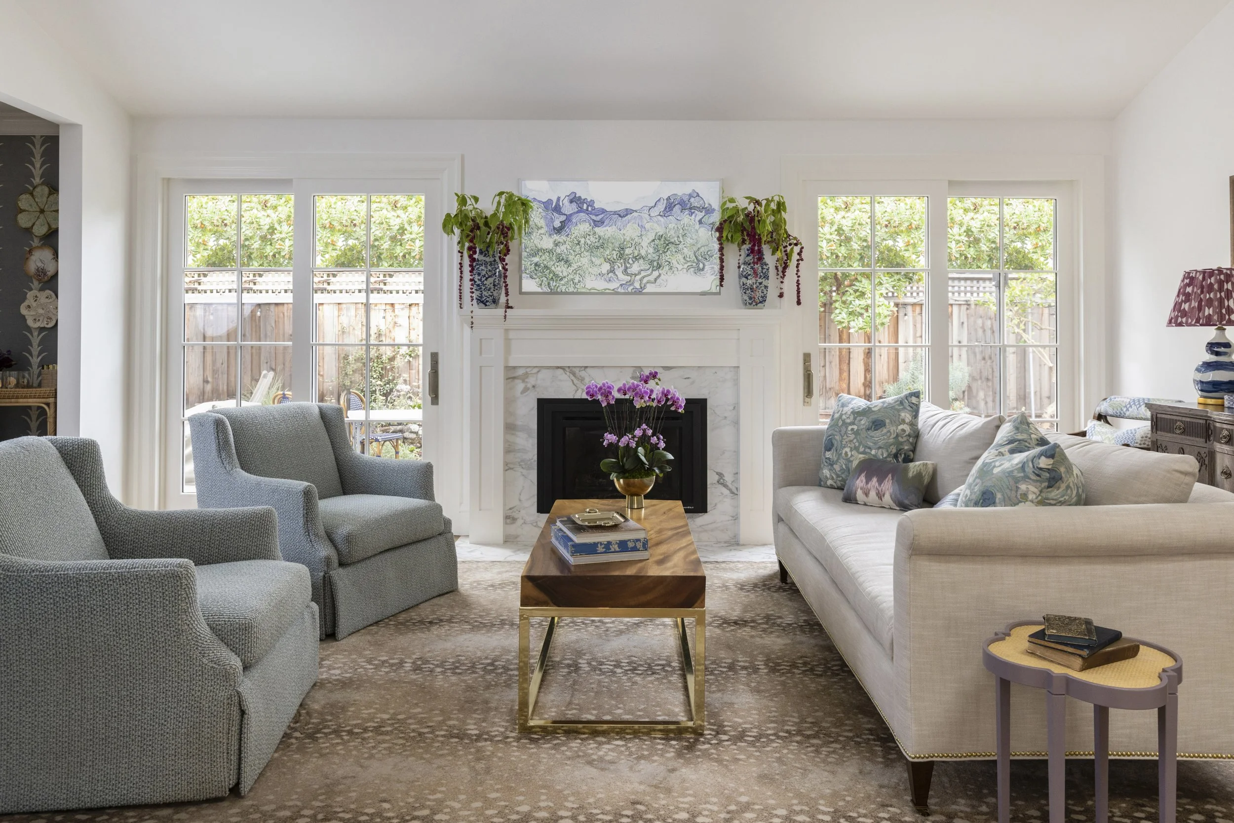 Living room with beige sofa, light blue armchairs, wooden coffee table with purple flowers, windows with garden view, fireplace, and artwork on the wall.