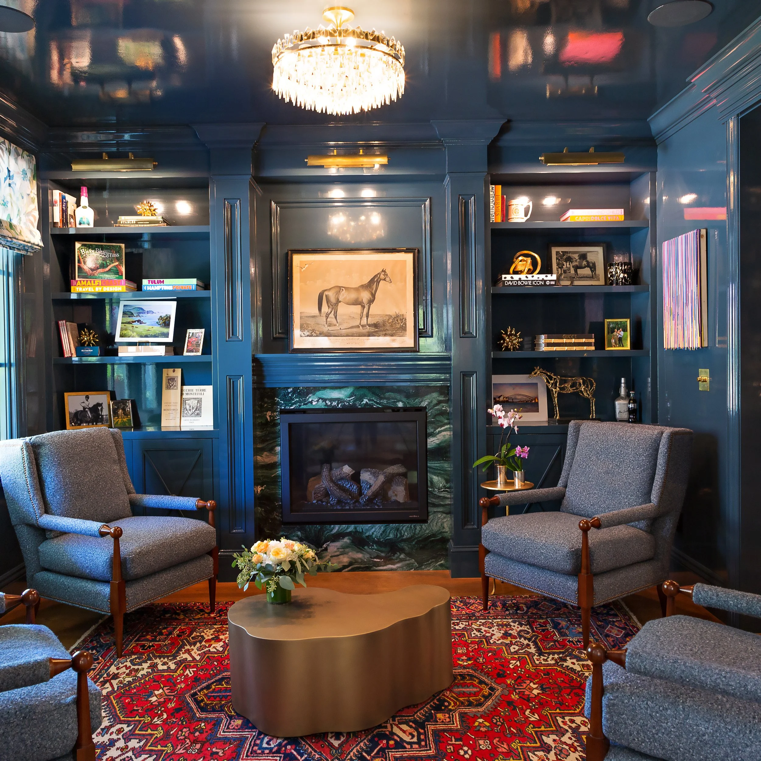 A cozy living room with blue paneled walls, a chandelier, and a fireplace with green marble. There are two gray armchairs around a gold coffee table with a flower arrangement. Built-in shelves with books, art, and decorative items surround the firepl