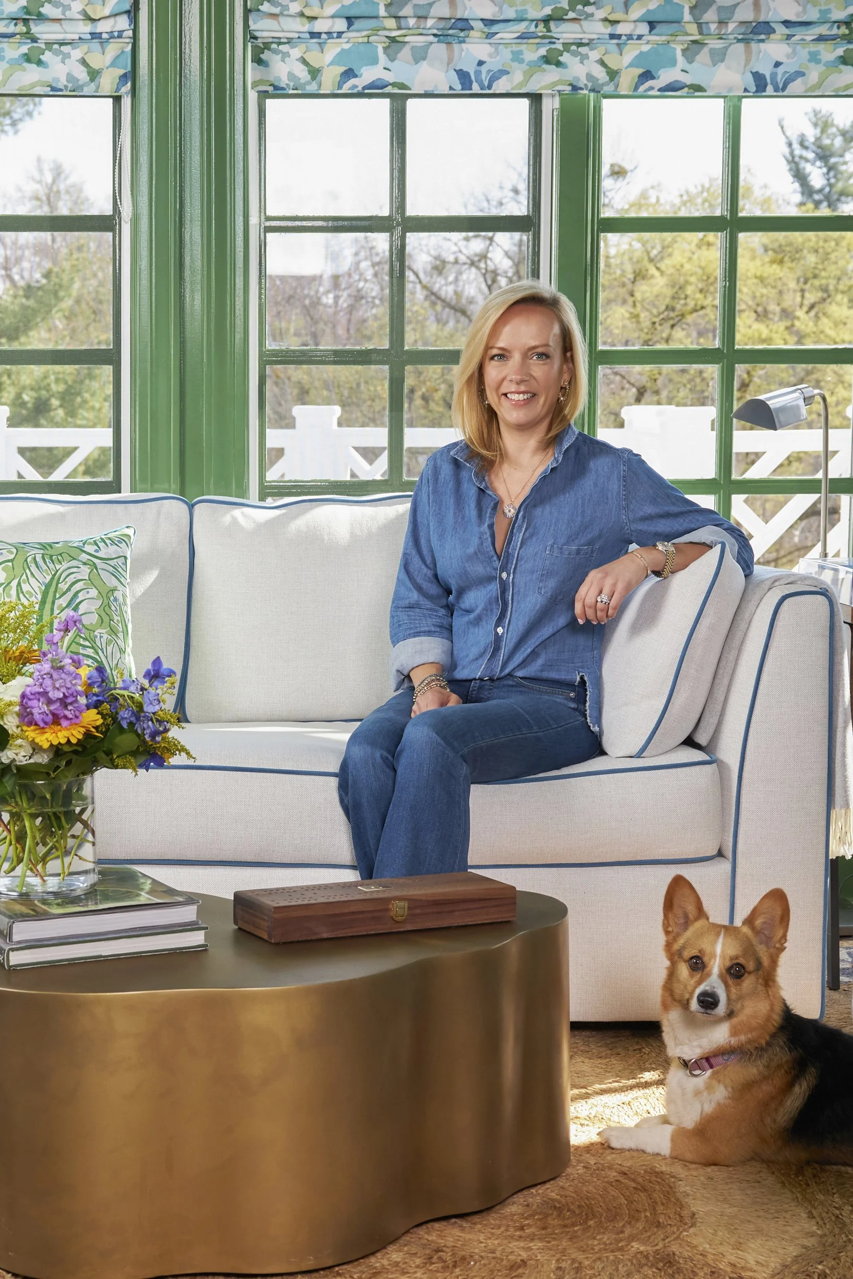 A woman sitting on a white sofa in a bright living room with large green-framed windows and trees outside. She has blonde hair, is smiling, and is wearing a blue denim shirt and jeans. A small dog is lying on the carpet in front of her, and there is a brass-colored coffee table with a vase of colorful flowers, a wooden box, and some books.