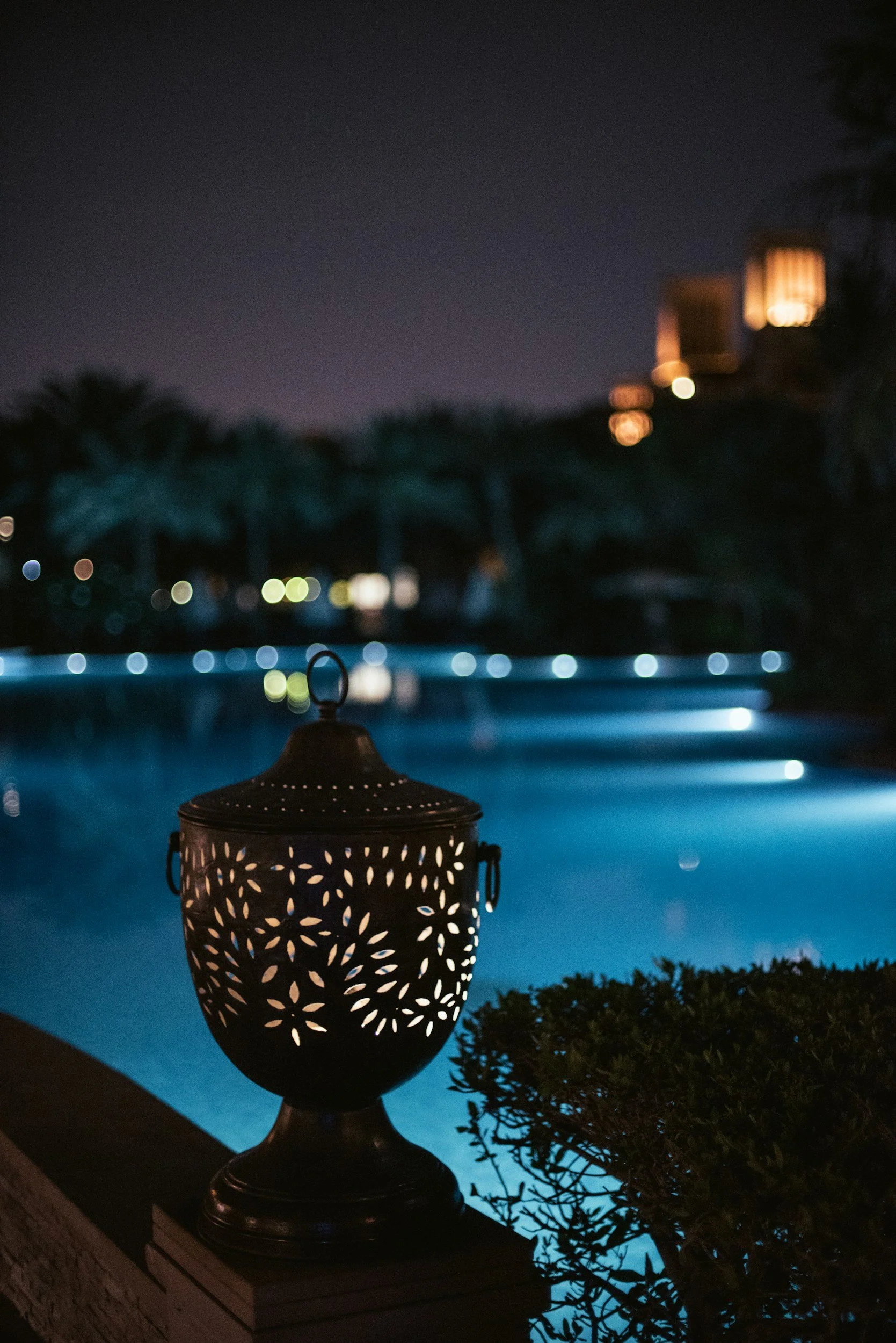 Decorative lantern by a pool at night, with illuminated water and city lights in the background.