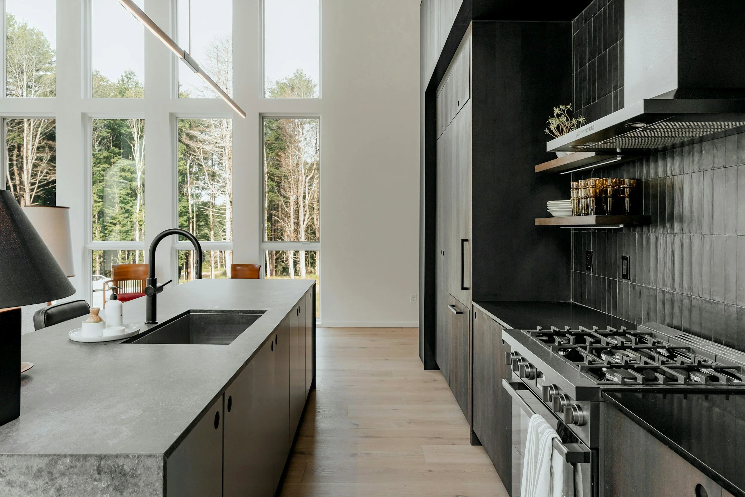 Modern kitchen with black cabinetry, black tile backsplash, stainless steel gas stove, black sink, large windows, wooden dining chairs, and light wood flooring.