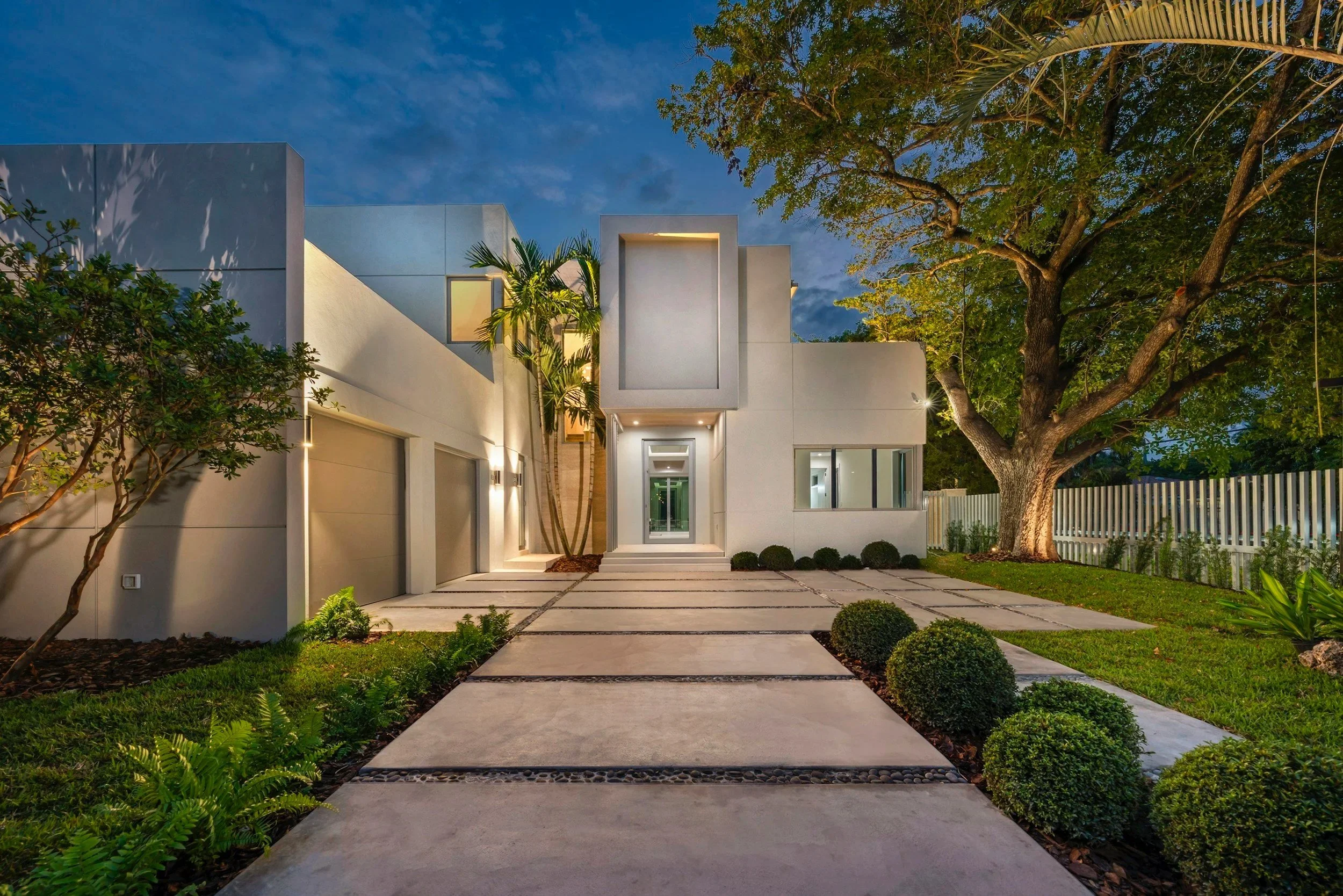 Modern white house at dusk with a paved walkway, surrounding greenery, and large trees in the yard.