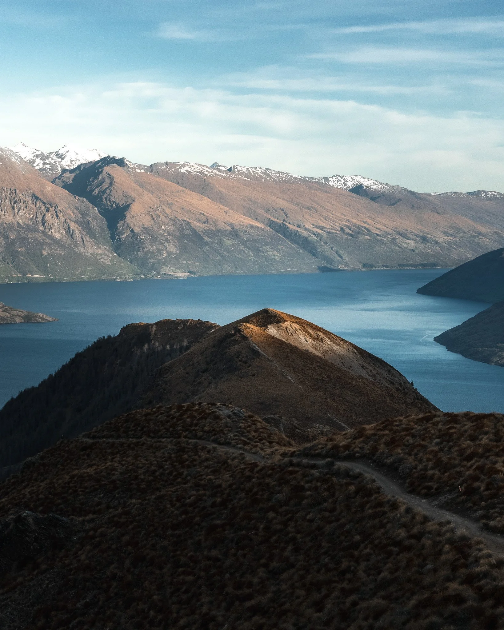 Mountain landscape with snow-capped peaks, brown and green slopes, a blue lake in the foreground, and a cloudy sky.