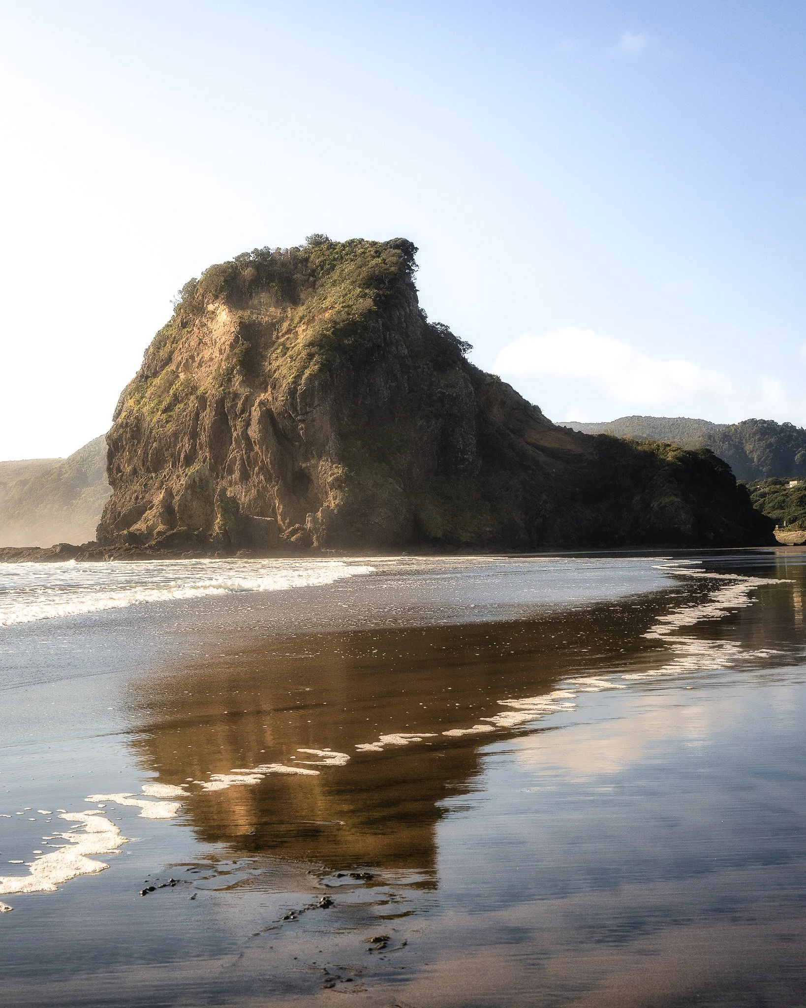 A large rocky cliff covered in greenery during sunset, with waves washing onto the sandy beach below and a partly cloudy sky overhead.