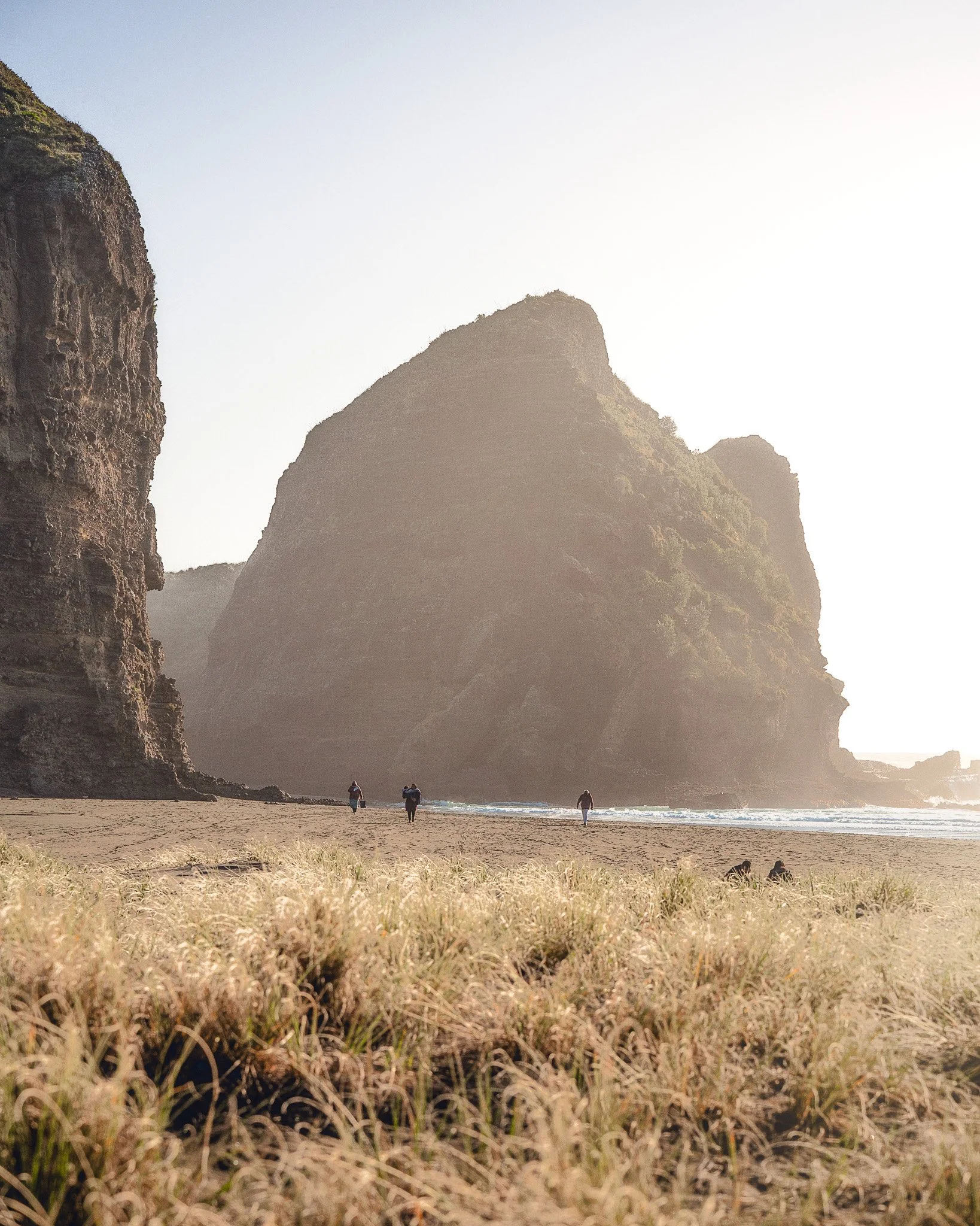 Beach with sand dunes in foreground, people walking along shoreline, large rocky cliffs in background, sunny day with clear sky.