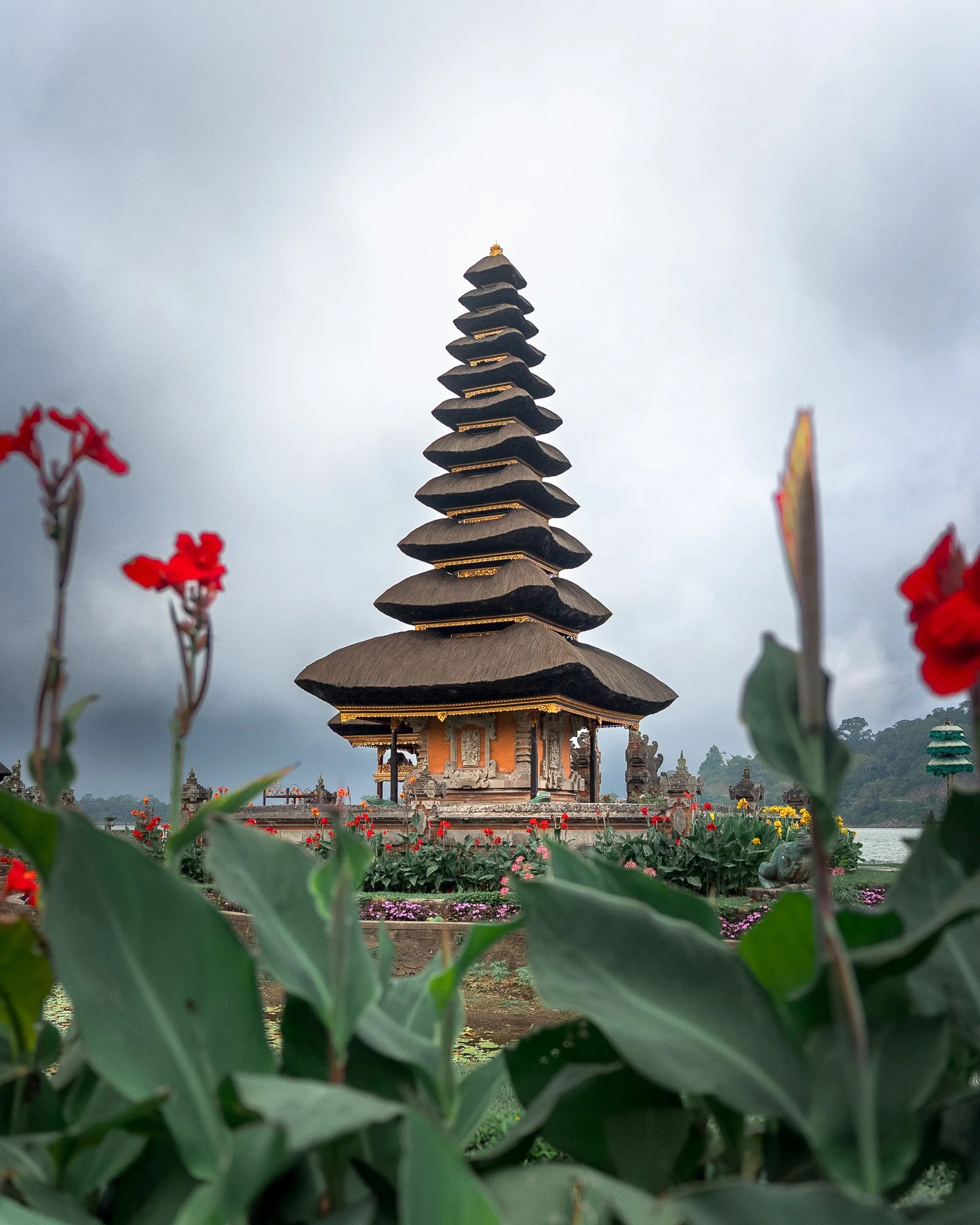 A multi-tiered pagoda with a dark, curved roof stands in a garden surrounded by colorful flowers. The sky is overcast with gray clouds.