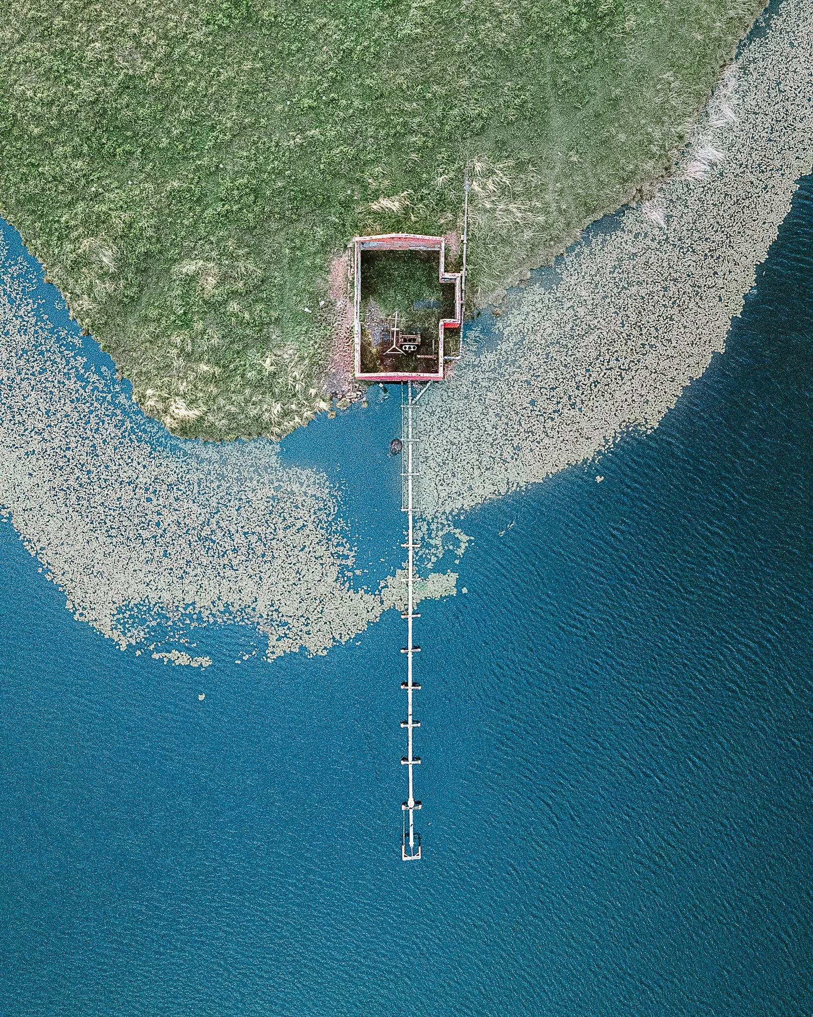 Aerial view of a house on a green island surrounded by water, with a long pier extending into the water. The house is located near the shoreline in a body of water, likely a lake or river, with trees on the island and a marina or dock area visible.