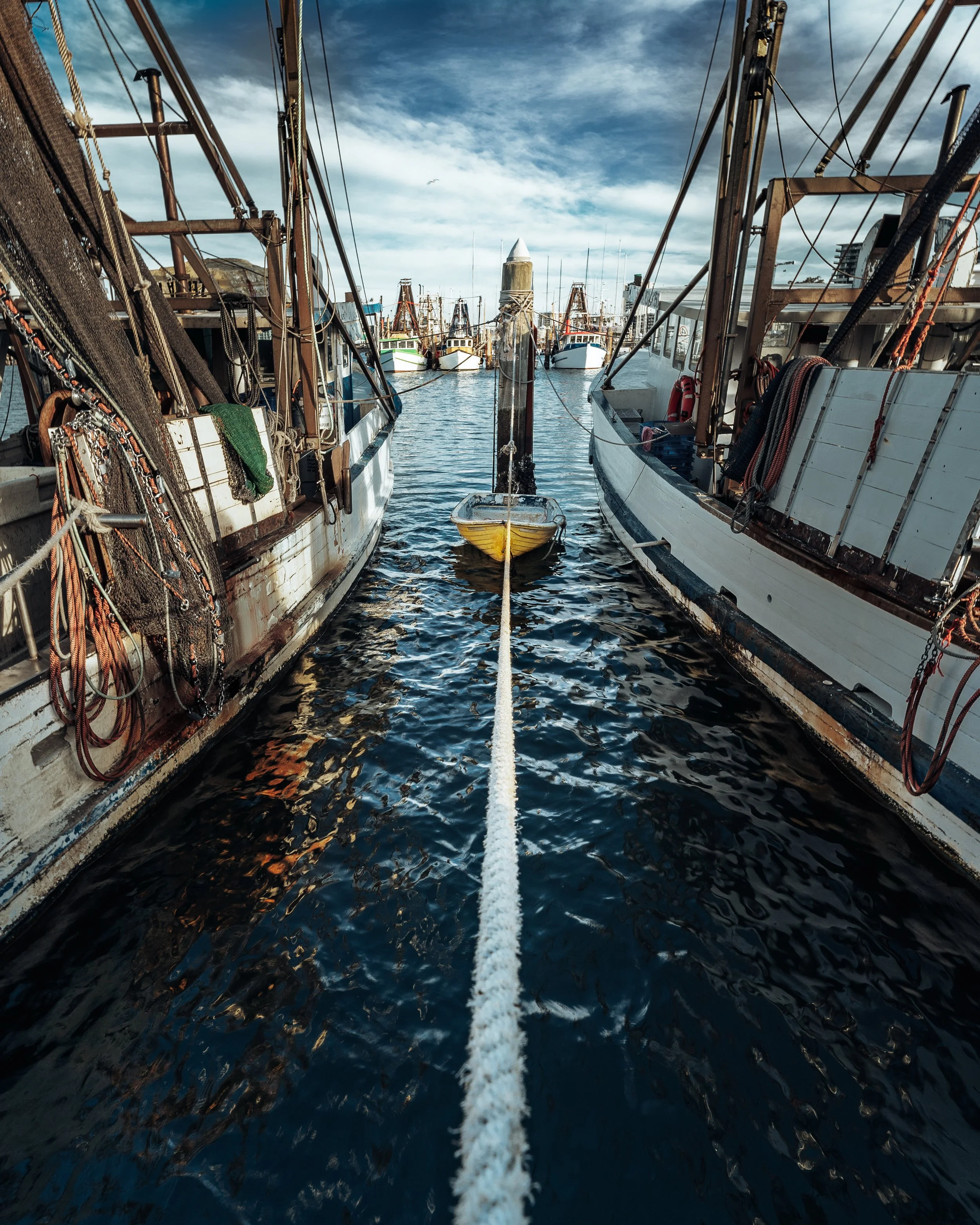 View between two boats at a harbor with a yellow rowboat tied to a central post, other boats in the background, and a partly cloudy sky.