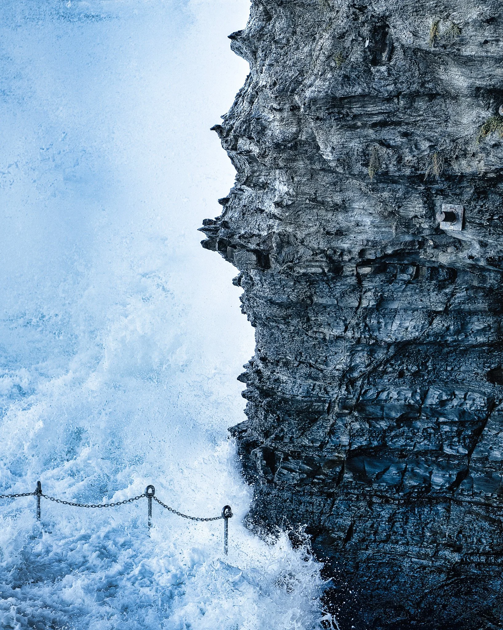 Close-up of a rocky cliffside with chains and posts visible at the bottom, overcast sky in the background.