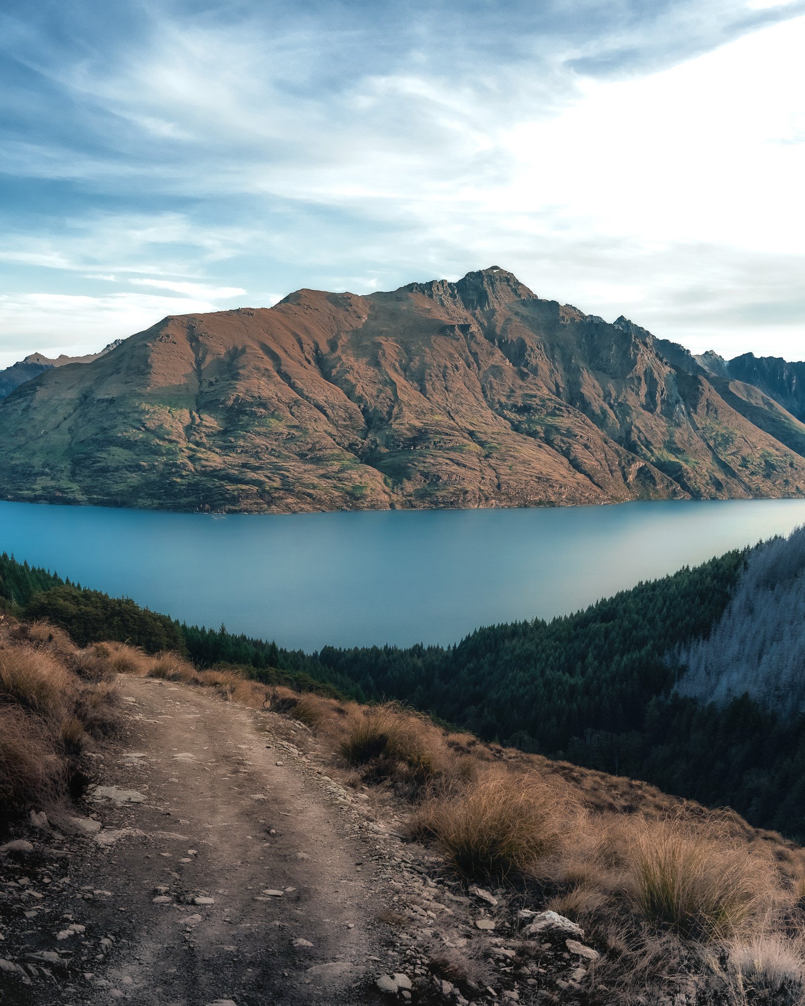 A trail through dry grass leading towards large mountain peaks with rugged, rocky slopes under a partly cloudy sky.