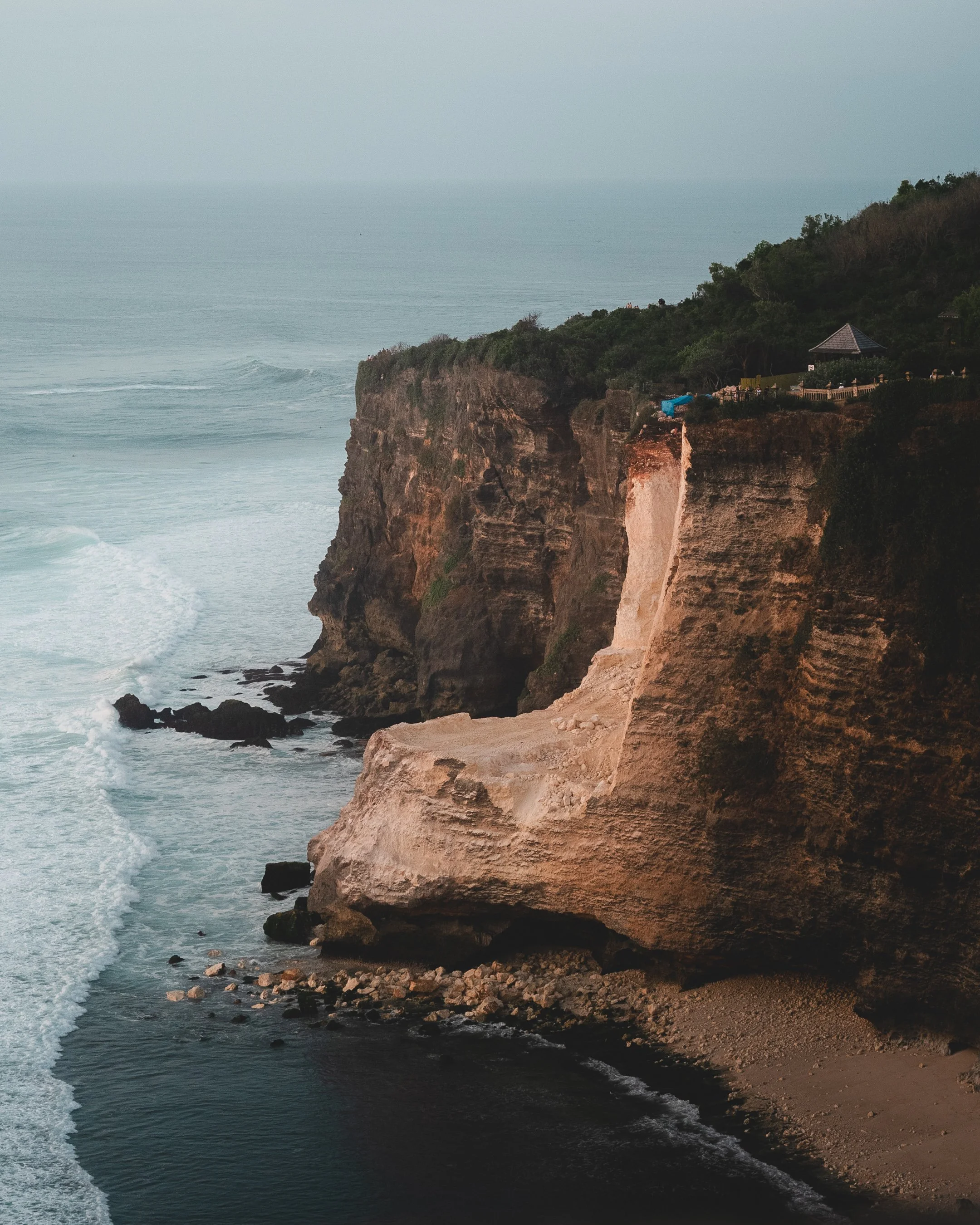 Cliffside with ocean waves crashing at the bottom, rugged rocky cliffs with green vegetation, and a small pavilion or gazebo on the top right.
