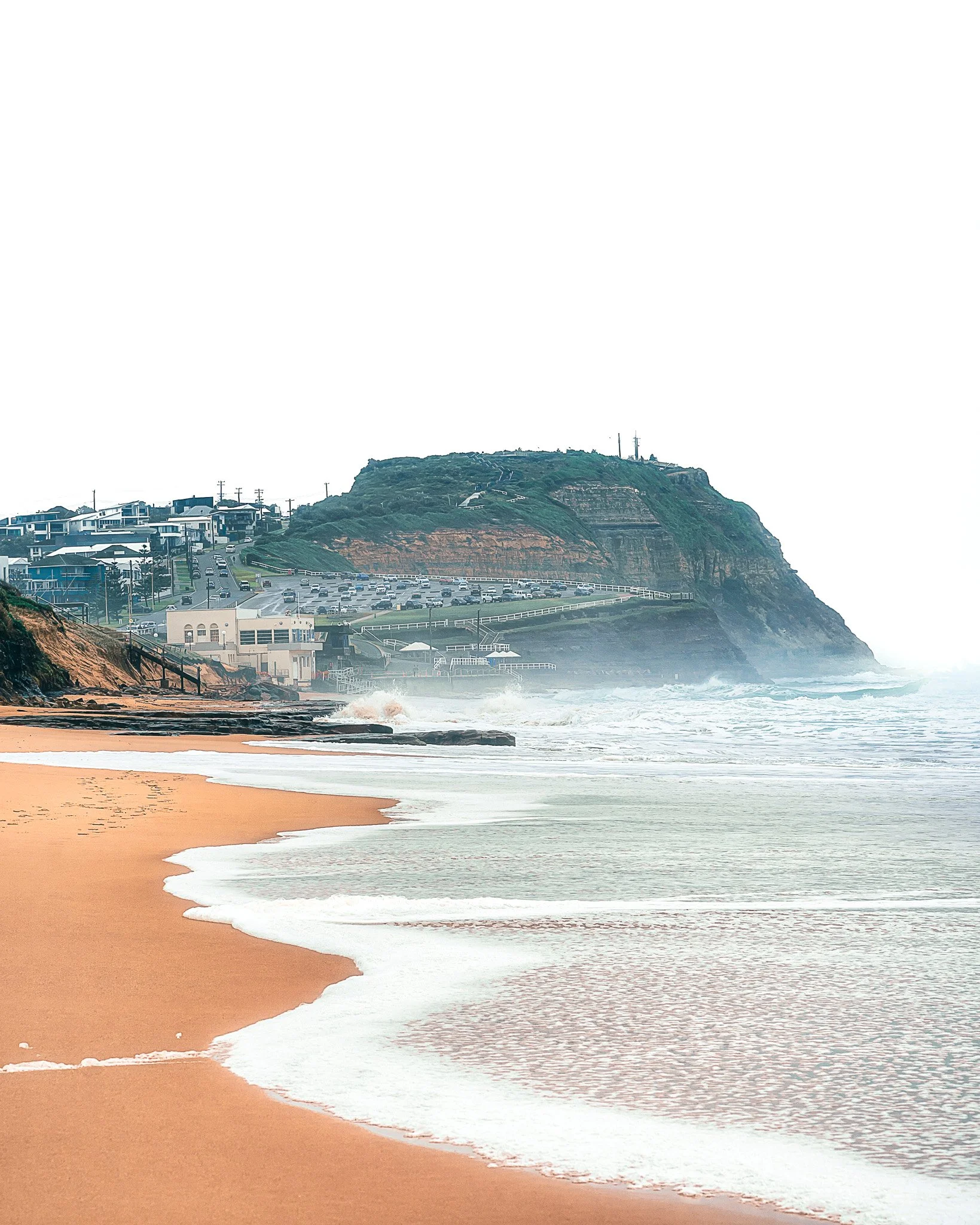 A sandy beach with gentle waves washing ashore, with rocky cliffs and residential buildings on top of the cliffs in the background, under a mostly overcast sky.