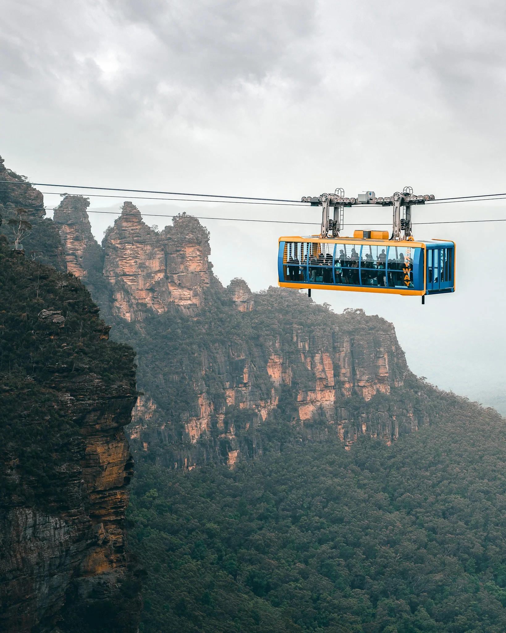 A cable car traveling over a mountainous landscape with lush green trees and rocky cliffs, and a cloudy sky overhead.