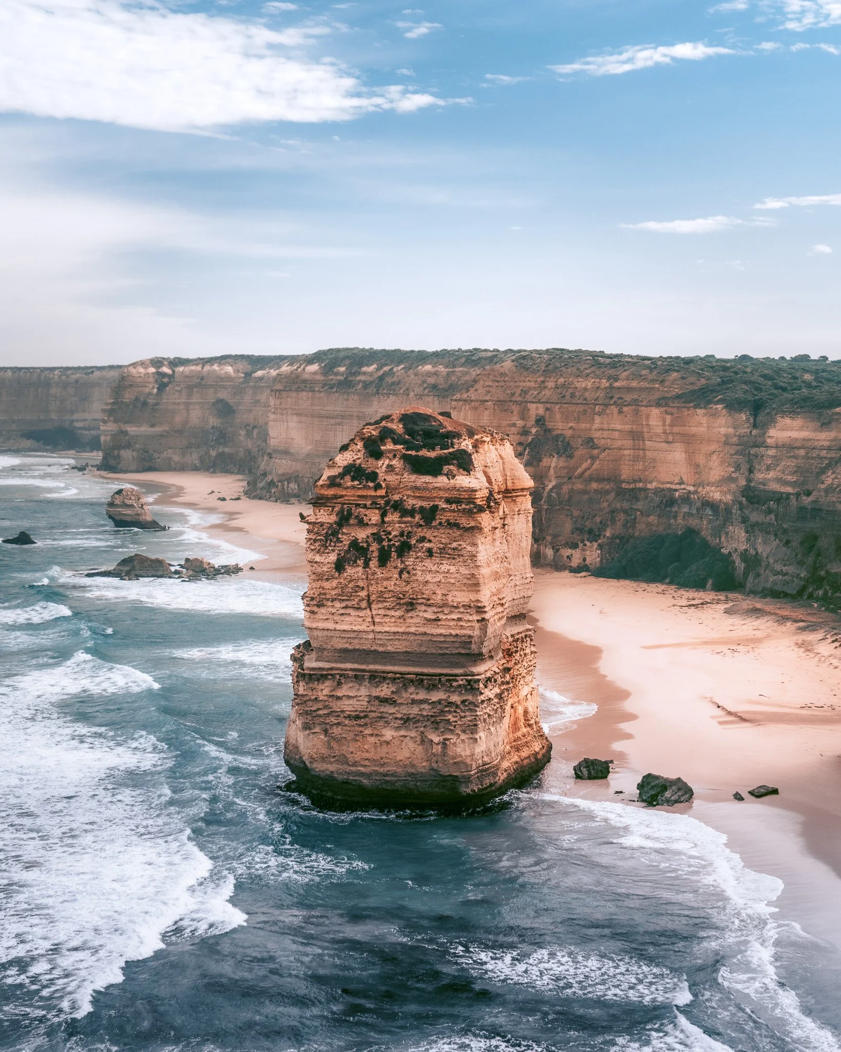 Cliffs and rock formations along a sandy beach, with ocean waves and blue sky with clouds