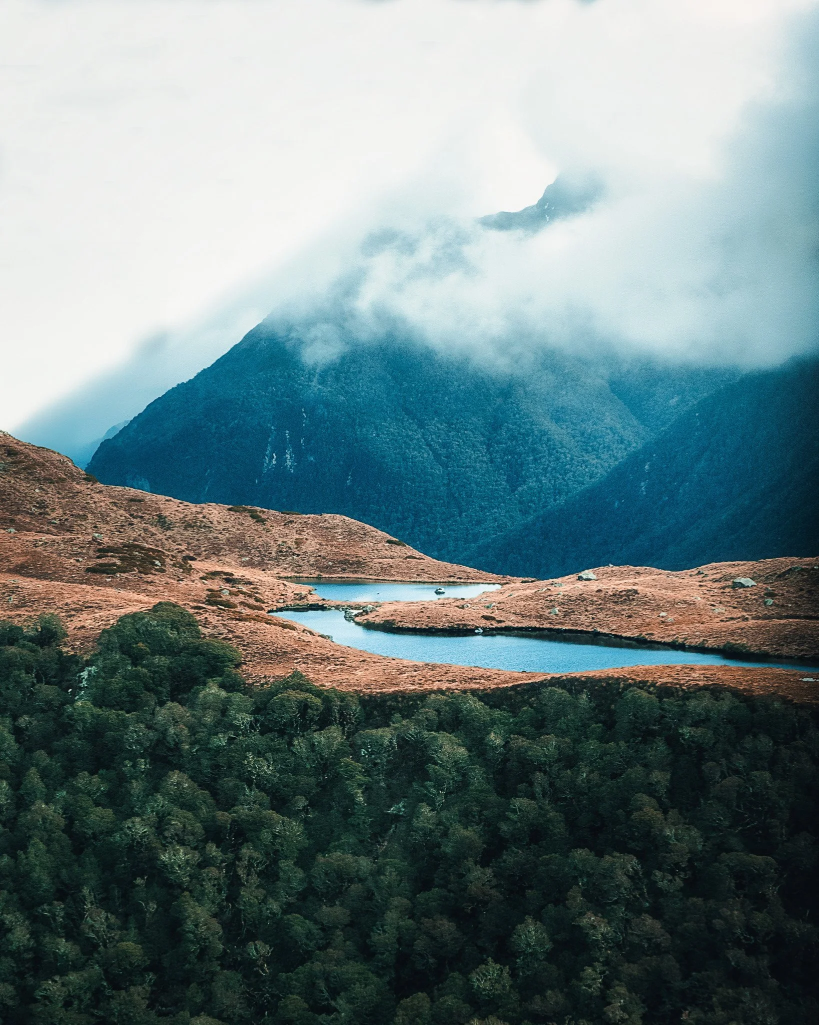 A mountain landscape with a steep, forested mountain partially shrouded in clouds, with a small lake and green forest in the foreground.
