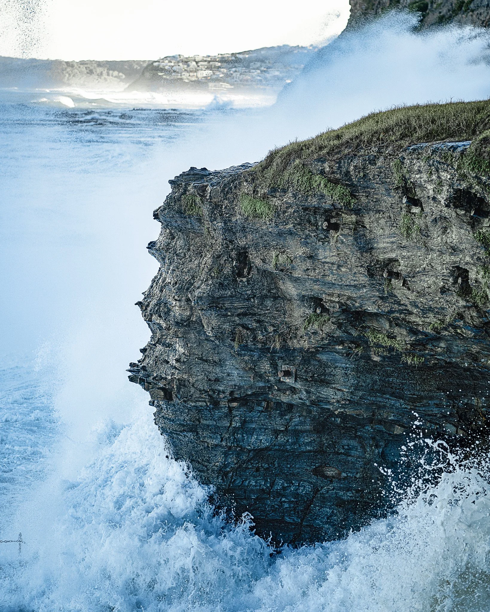 Ocean waves crashing against rocky cliffside with a coastal town visible in the distance on a clear day.