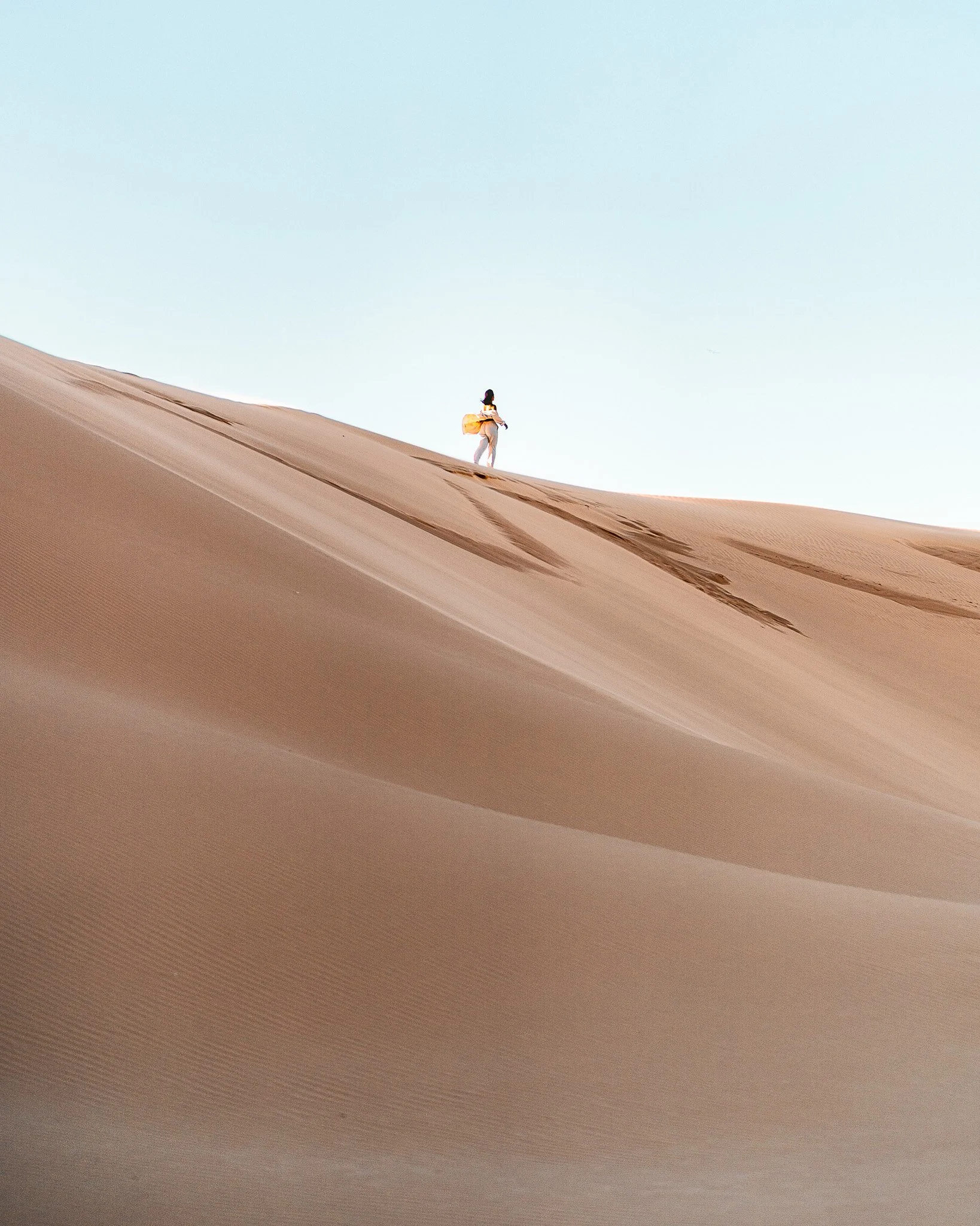 A person with a backpack walking on a sand dune in a desert landscape under a clear sky.