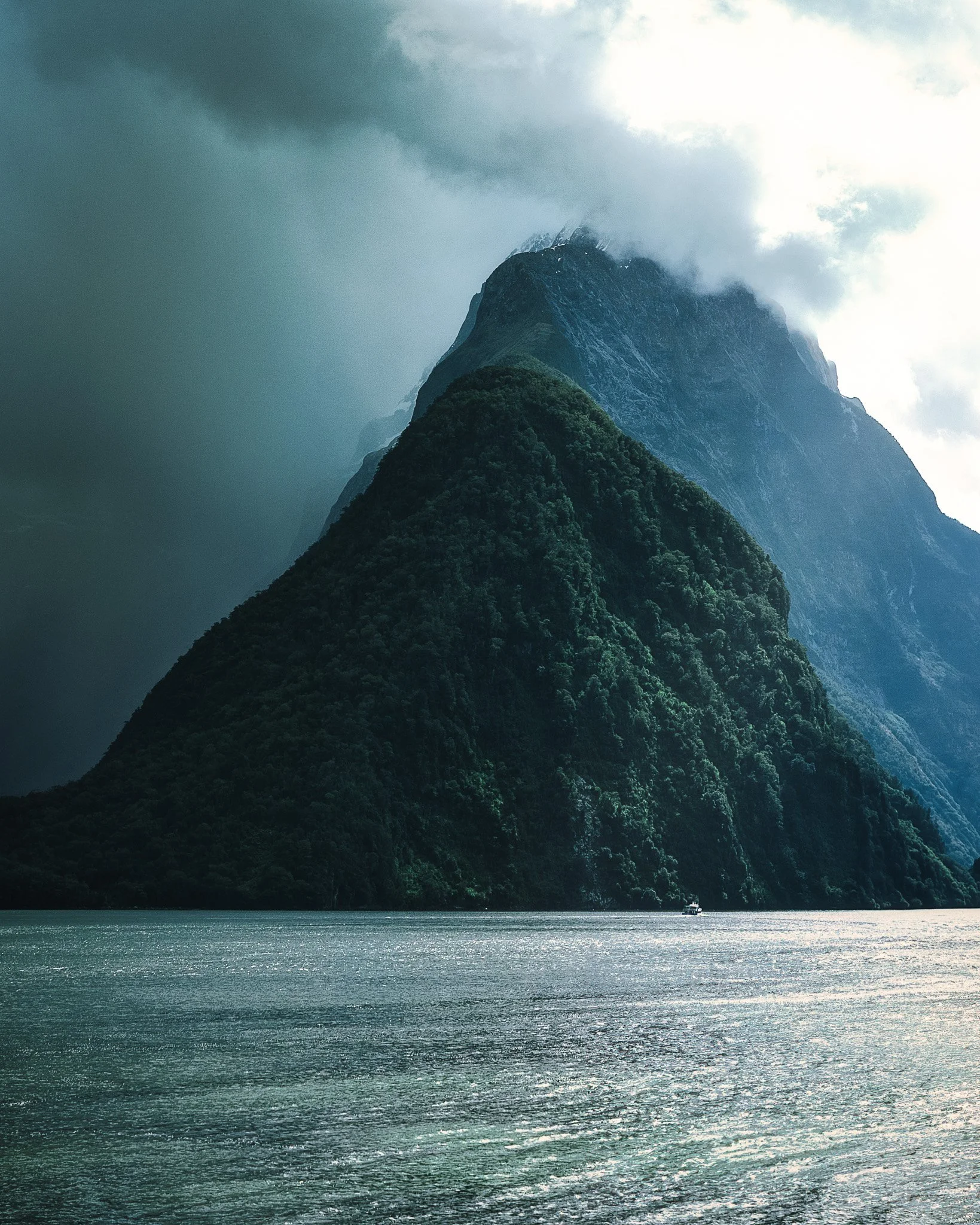 A mountain with snow and fog at the top, surrounded by green forest, near a body of water with a boat.