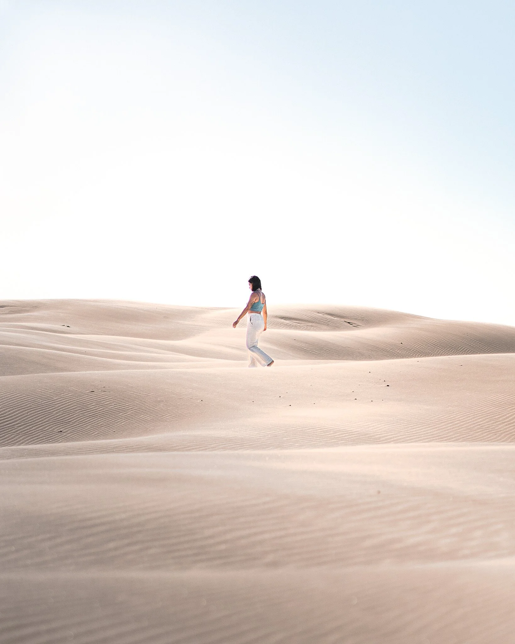 Woman walking in a vast sandy desert with sand dunes and a pale blue sky.
