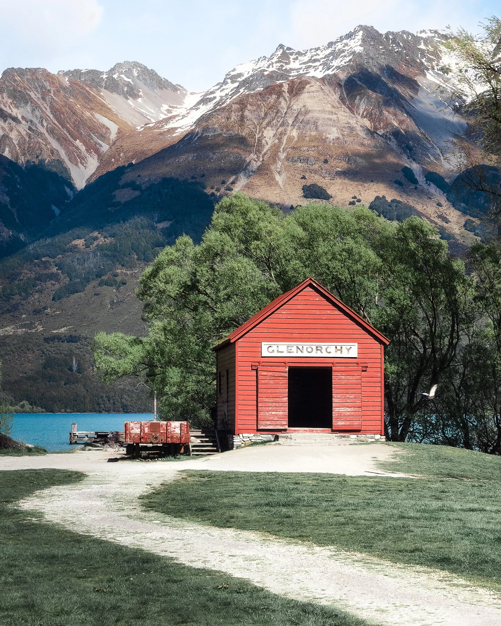 A red wooden building with a sign that says 'Glenorchy' surrounded by green trees, with a mountain range with snow patches in the background and a body of water to the left.