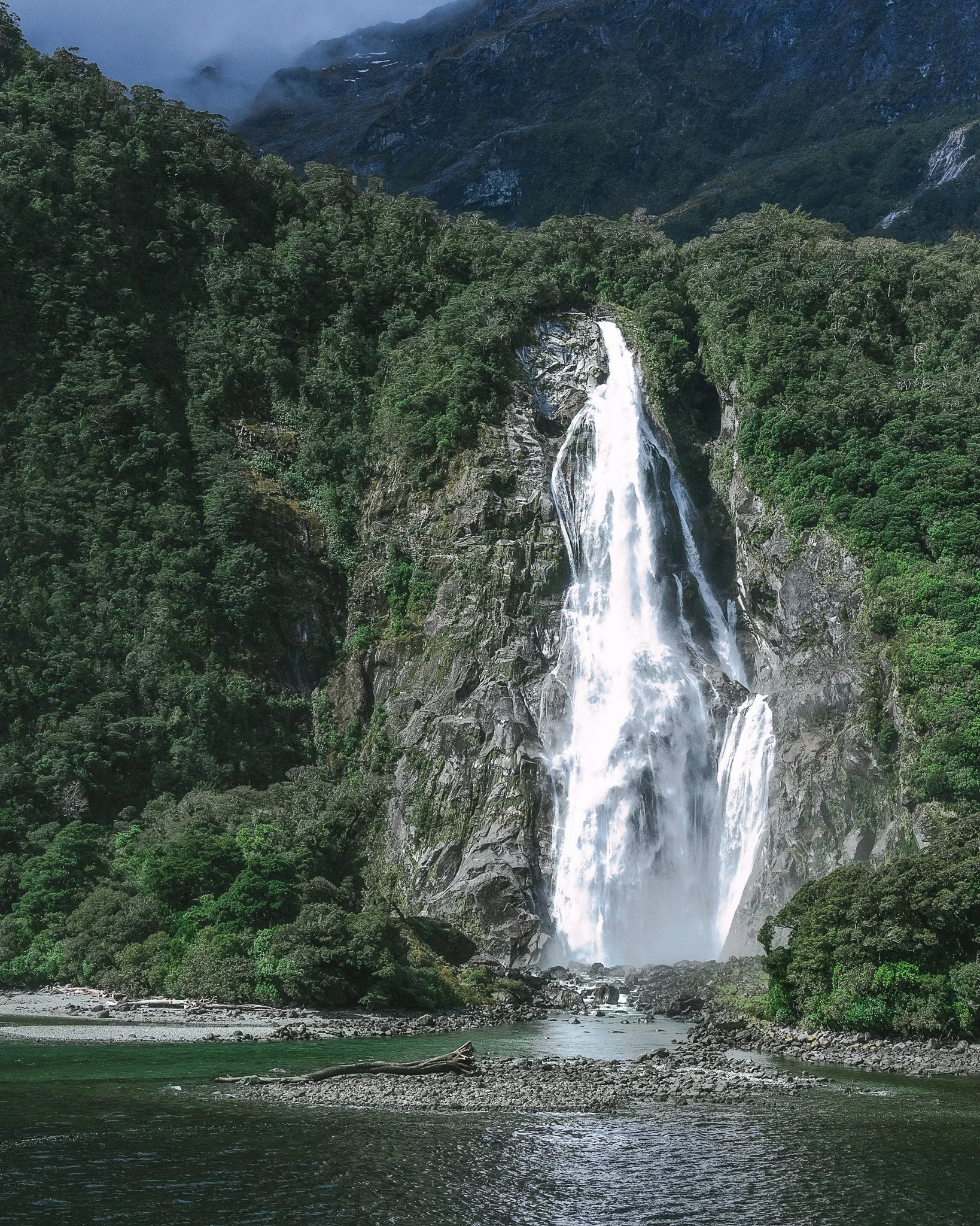 A lush green forest with a tall waterfall cascading down a rocky mountainside into a river below, surrounded by misty mountains in the background.