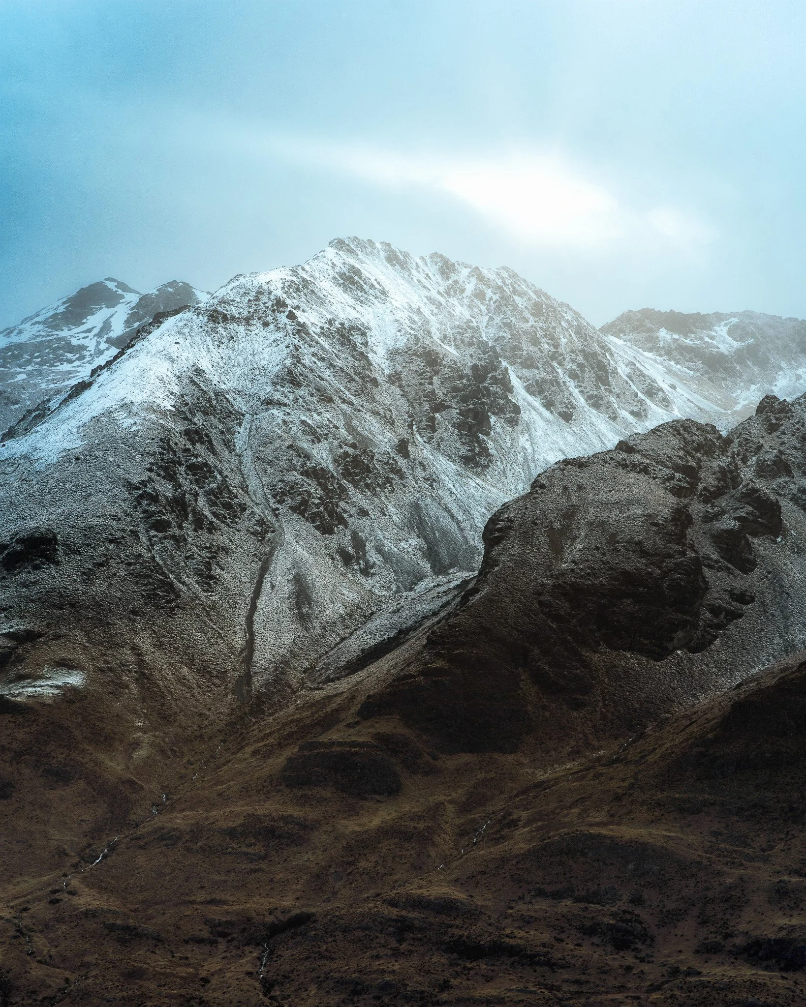 Snow-capped mountain peaks against a cloudy sky with dark rocky foreground.