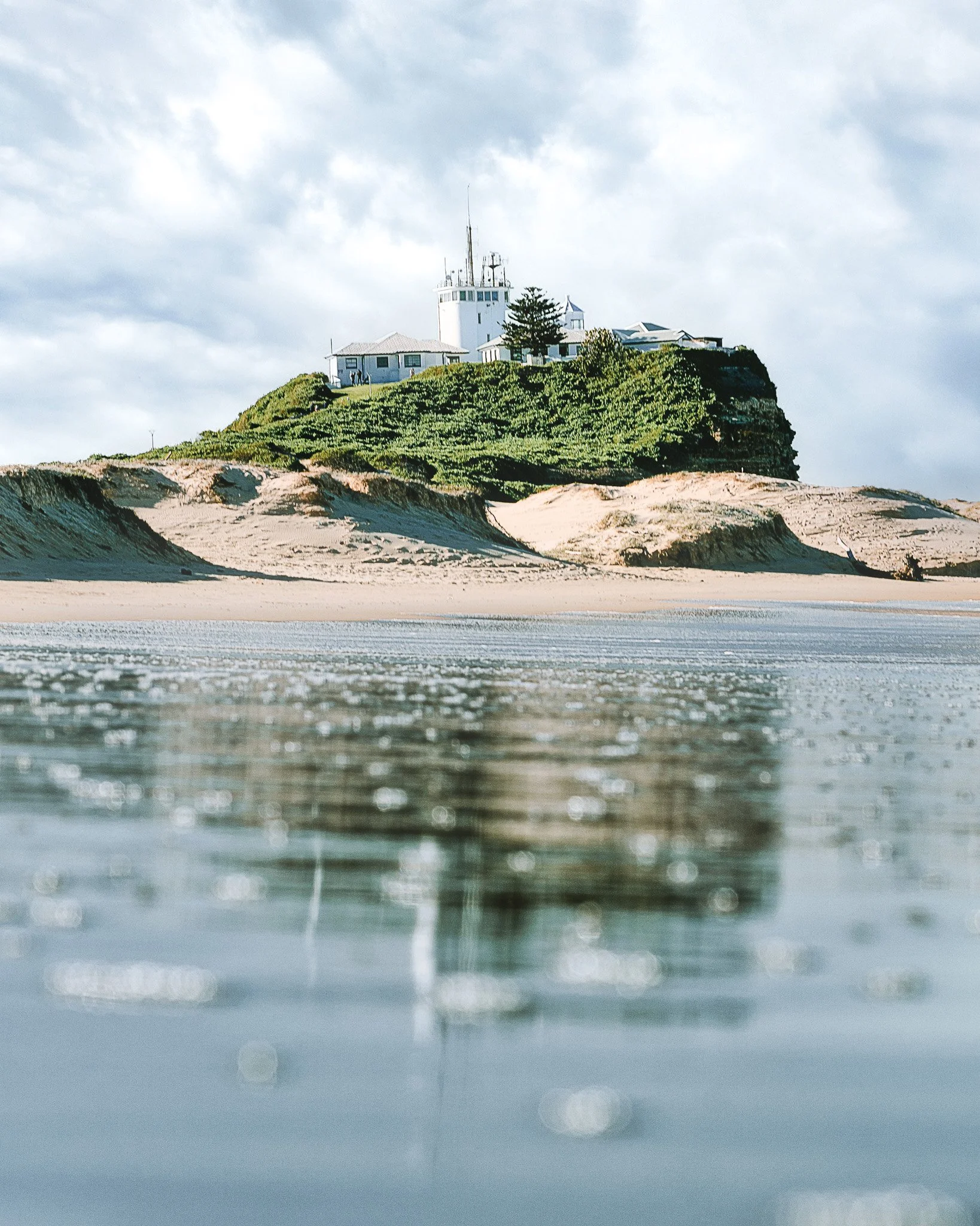 View of a lighthouse on a hill with greenery and buildings, seen from near the shoreline with waves in the foreground.