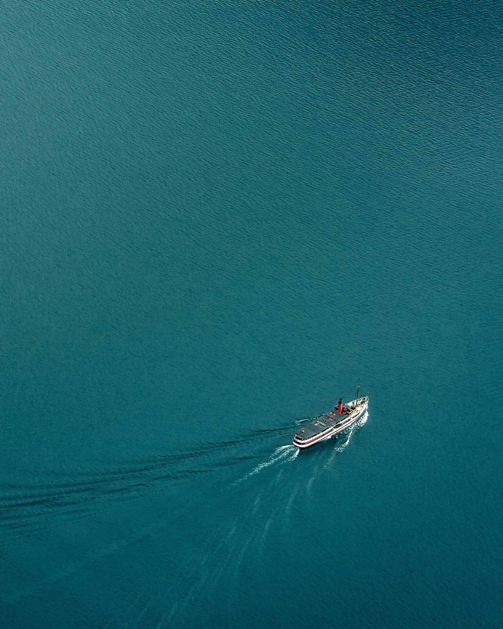Aerial view of a vintage steamship sailing on calm blue water with a dotted wake behind it.