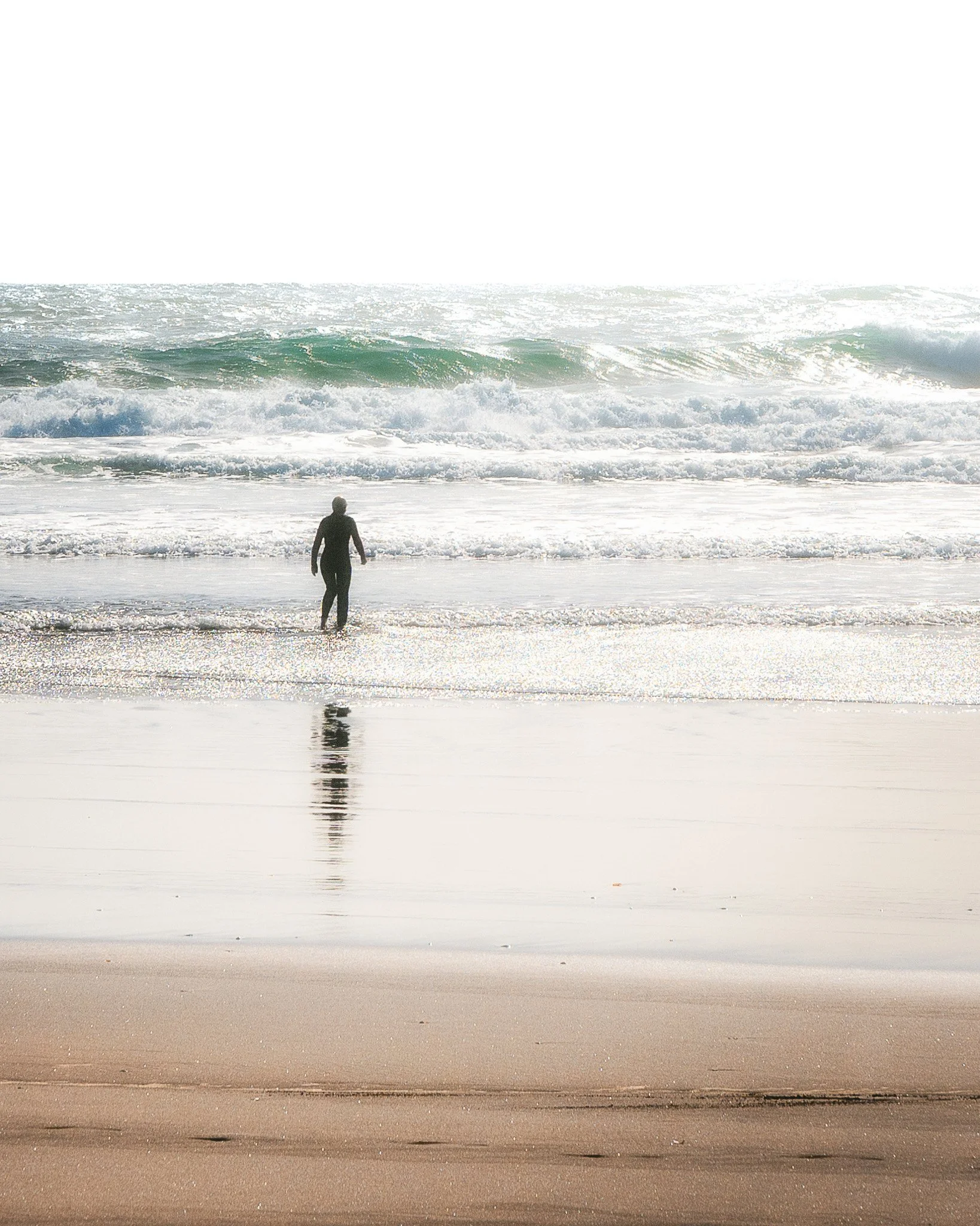 A person walking on the wet sandy beach with ocean waves crashing nearby under a bright, sunny sky.