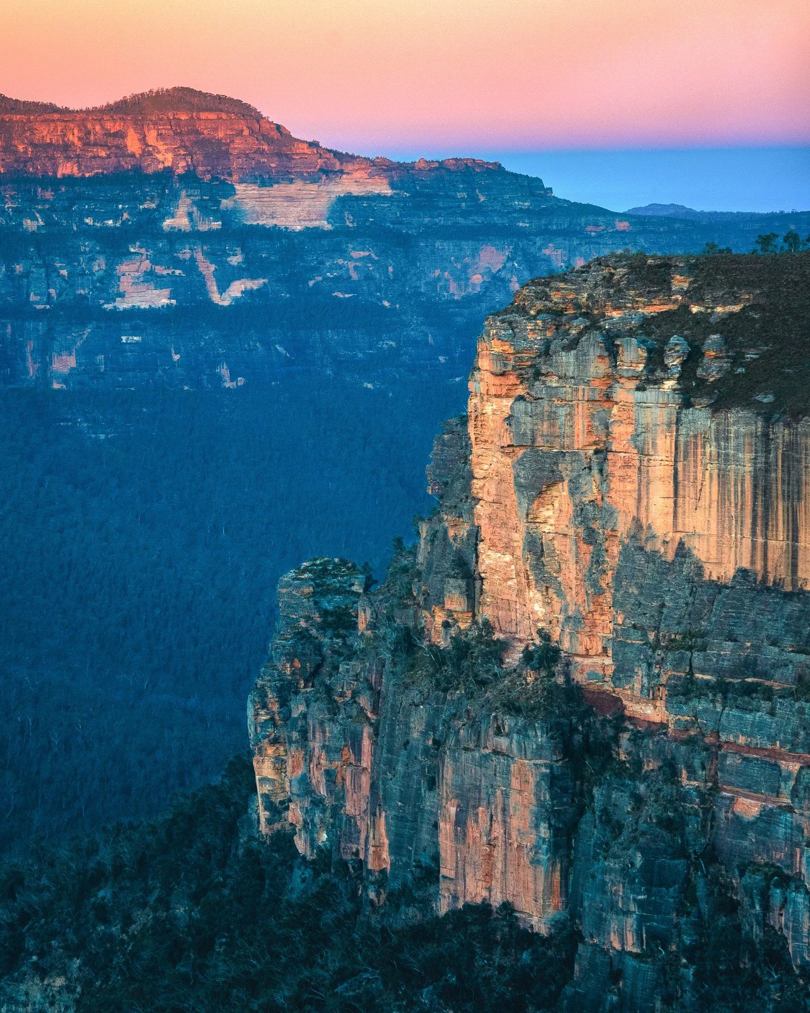 Sunset over a canyon with layered rock formations and trees, illuminated with soft pink, purple, and orange colors.