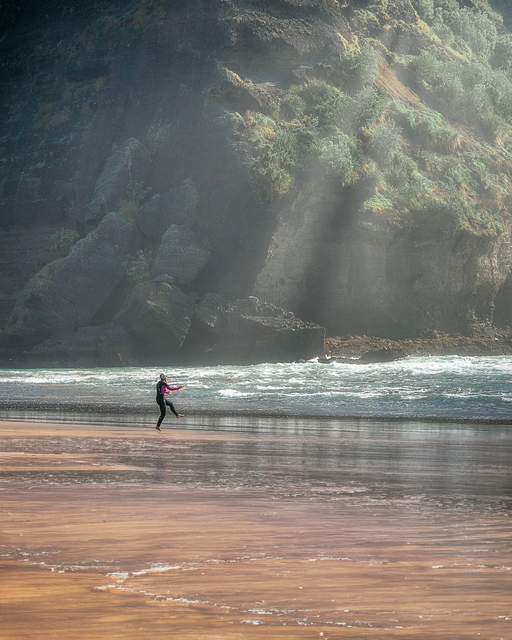 A person in a pink and black jacket flying a kite on a beach with large rock formations and green vegetation in the background.