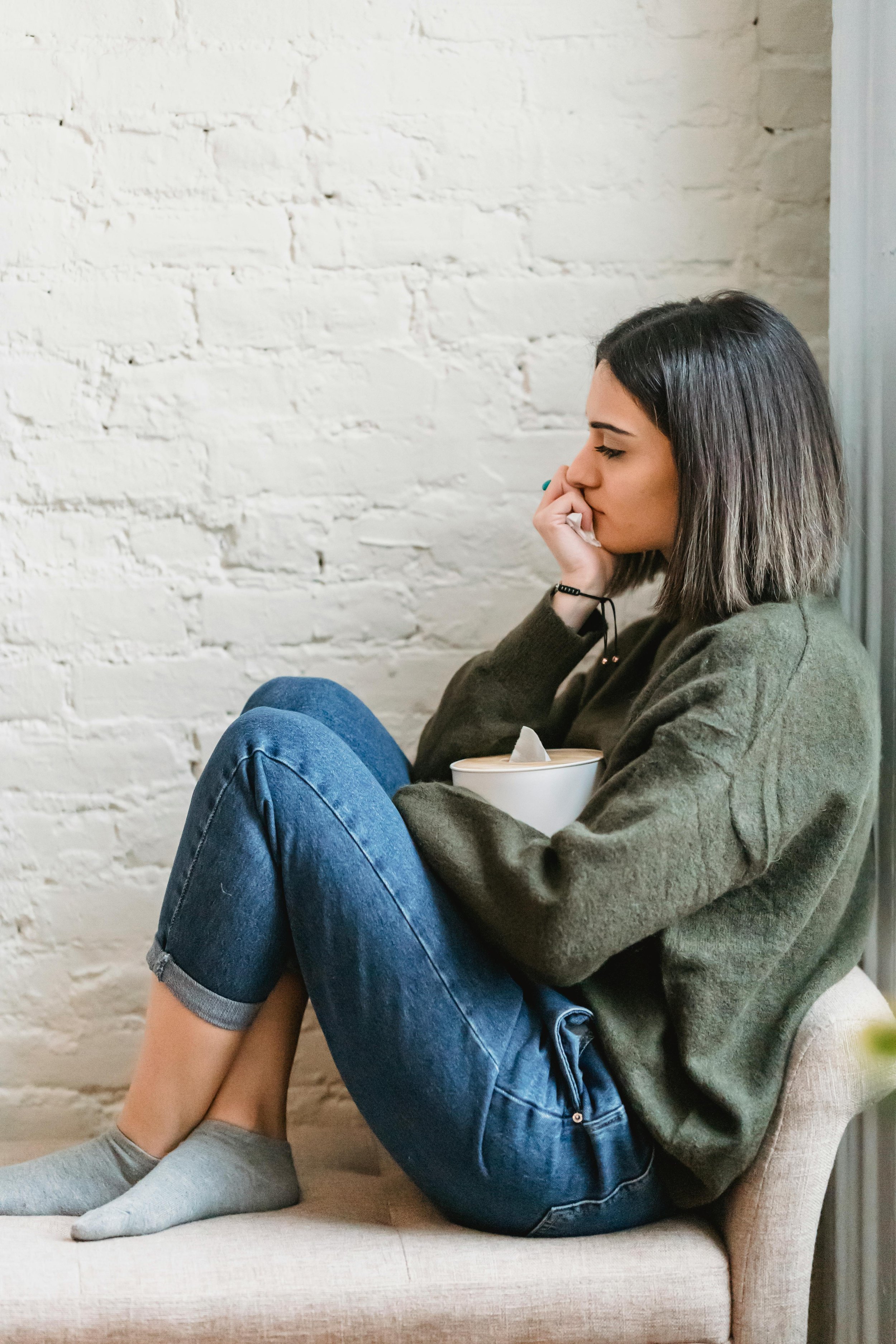 A young woman sitting on a beige bench with a white brick wall in the background, holding a bowl and appearing to be sad or contemplative.