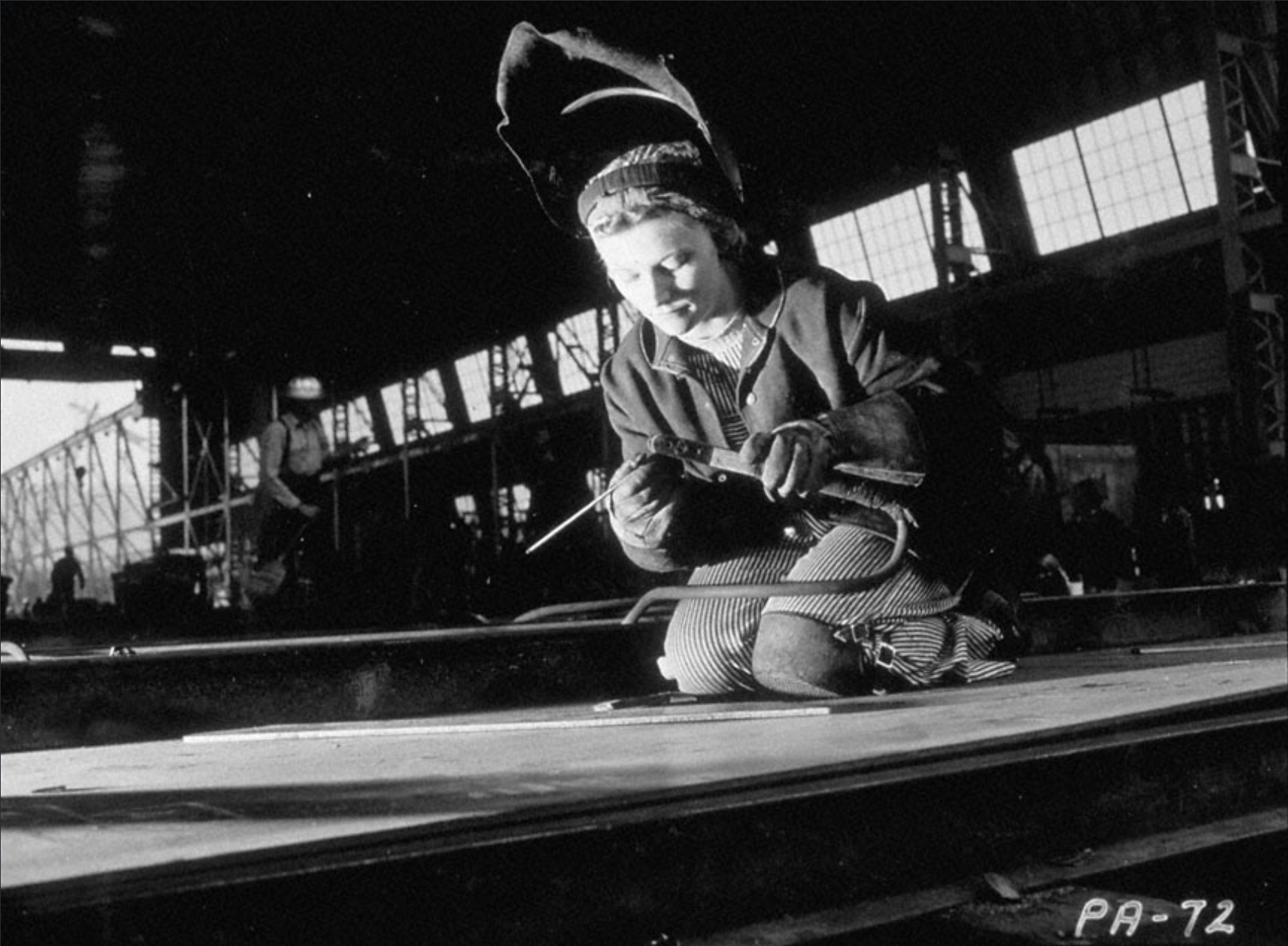 Black and white archival photo: A woman kneeling while welding at a World War II shipyard facility.