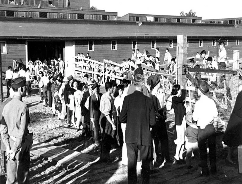 Black and white archival photo: People of Japanese descent moving through an "assembly center."