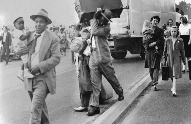 Black and white archival photo: People carrying children and luggage while evacuating.