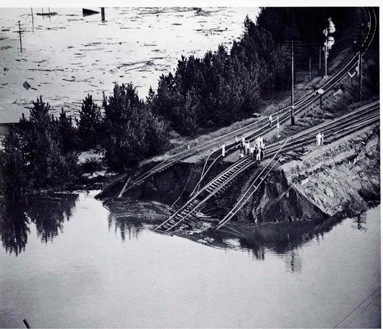 Black and white archival photo: A broken levee surrounded by water. People standing on top of the levee survey damaged railroad tracks and flooding.