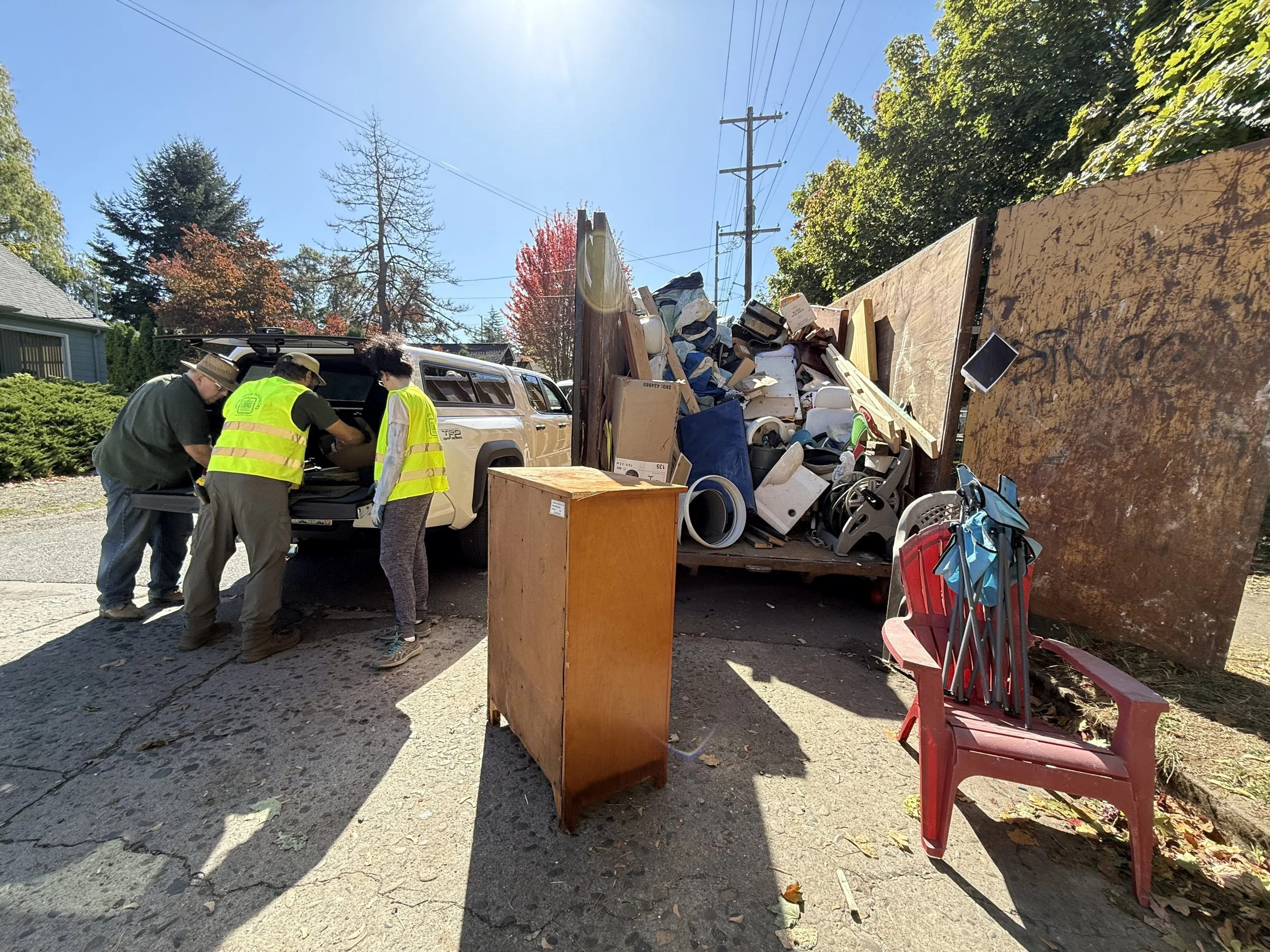 Three people unload furniture and other items into a dumpster.