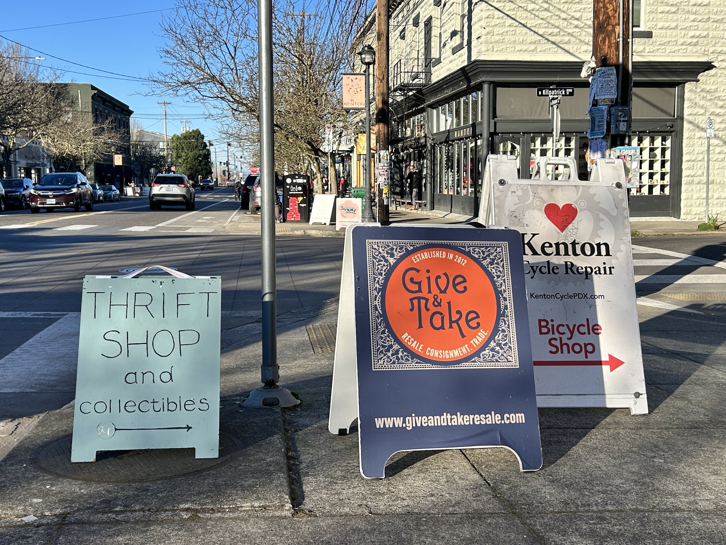 Three A-frame signs on a commercial district corner promote thrift stores and a bicycle repair shop.