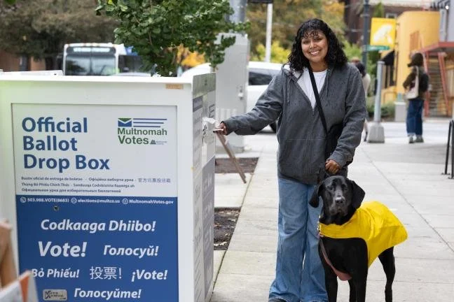 A person with a dog smiles while depositing a ballot into an official ballot drop box.