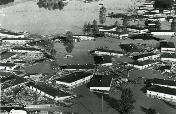 Black and white archival photo: Aerial view of a flooded neighborhood; only the top floor of buildings and treetops are visible.