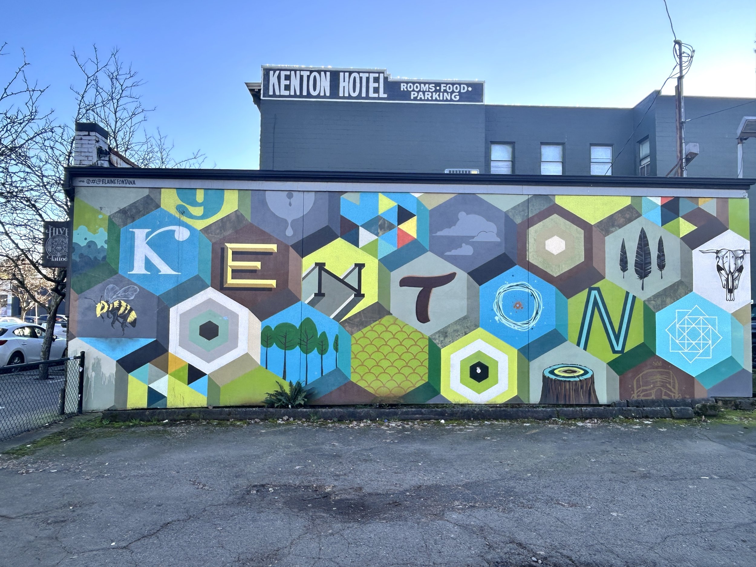 Colorful mural consisting of hexagon shapes, each featuring a unique design or object; six hexagons feature one letter each, spelling "KENTON". In the background, the top floor sign for the Kenton Hotel is visible.