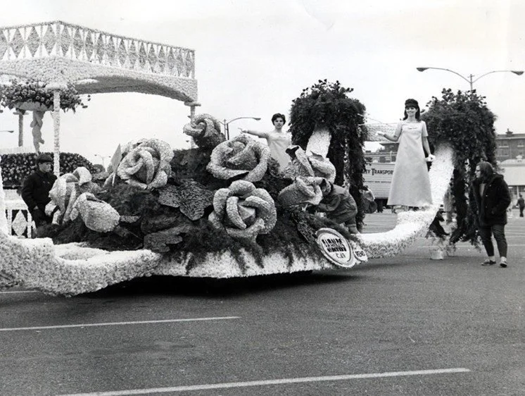 Black and white archival photo: Women standing on a parade float decorated with larger-than-life roses.