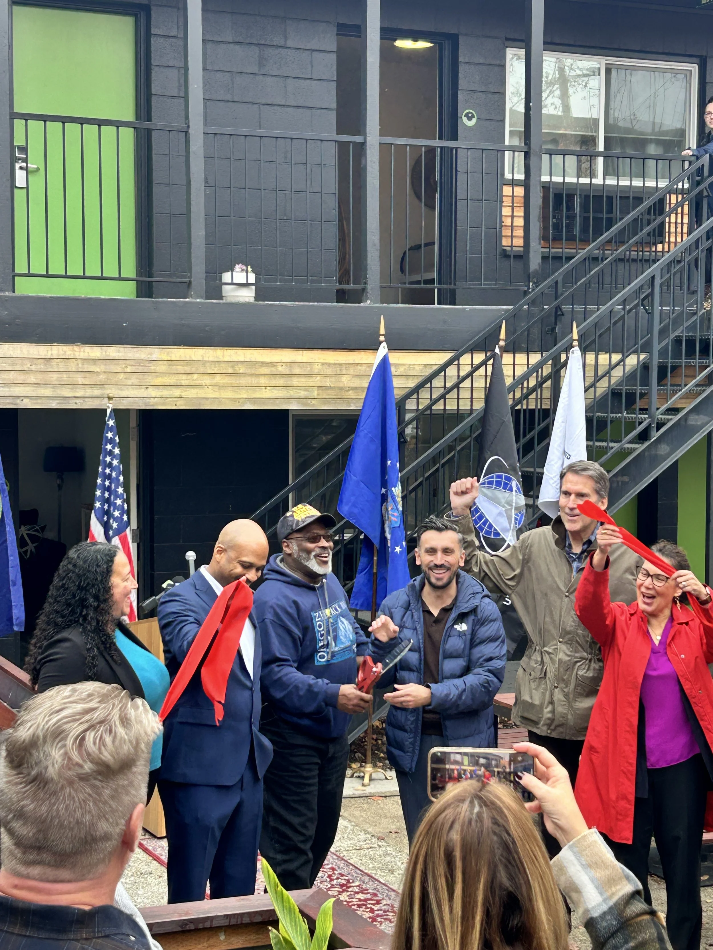 Six people excitedly cut a big red ceremonial ribbon while standing in front of a shelter facility.