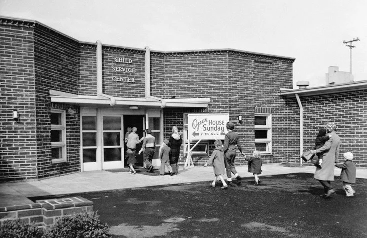 Black and white archival photo: Parents bringing their children into a brick building.