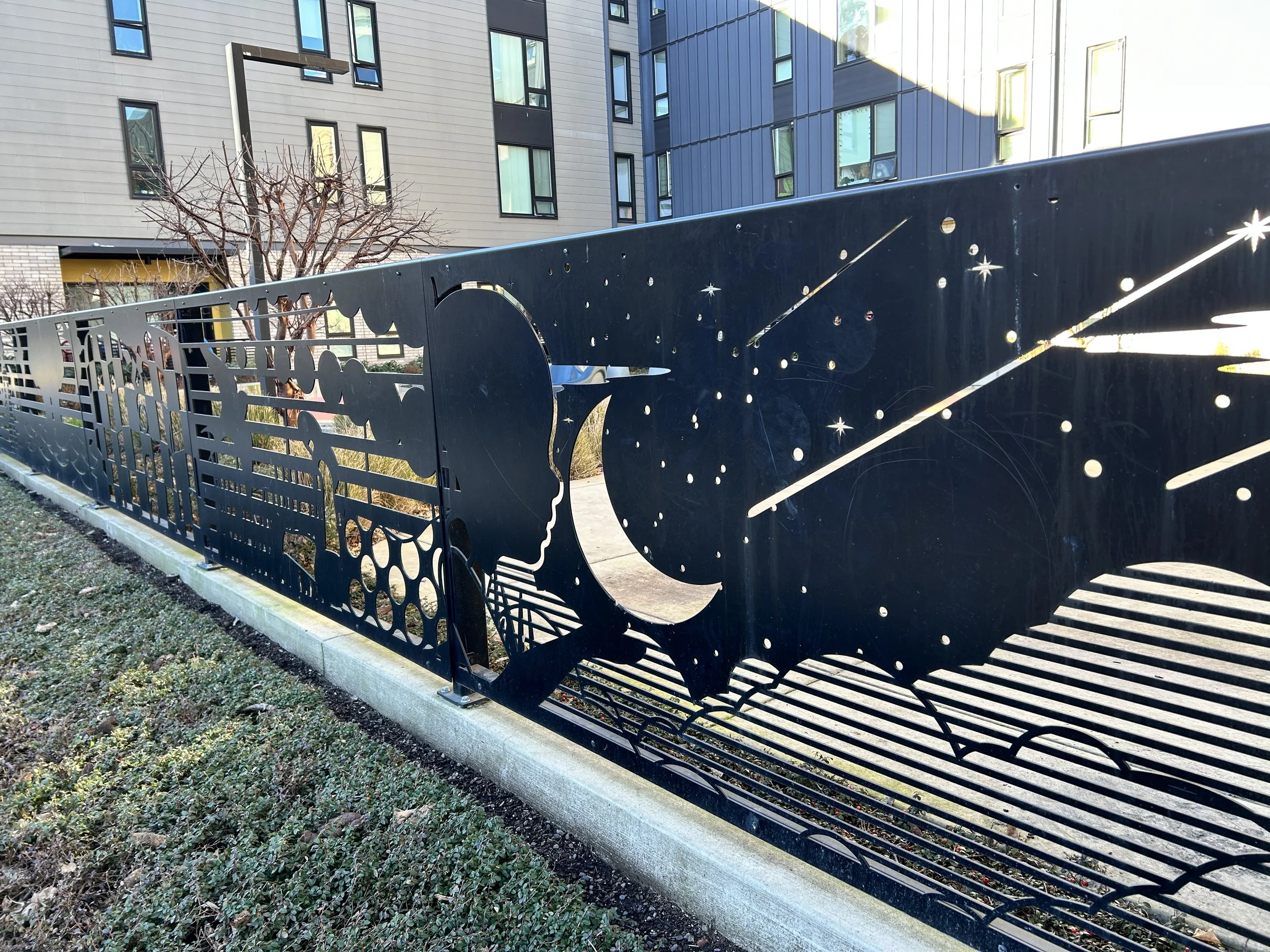 Decorative black fence featuring the silhouette of a child with hills, the moon, and stars through the use of cutouts; apartment buildings are visible in the background.