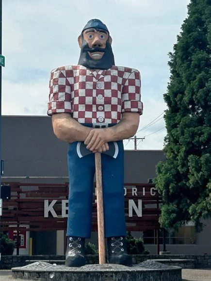Paul Bunyan statue depicts the folk character wearing a red and white checkered button up shirt, blue jeans, and boots, with his hands resting on a double sided ax standing upright.