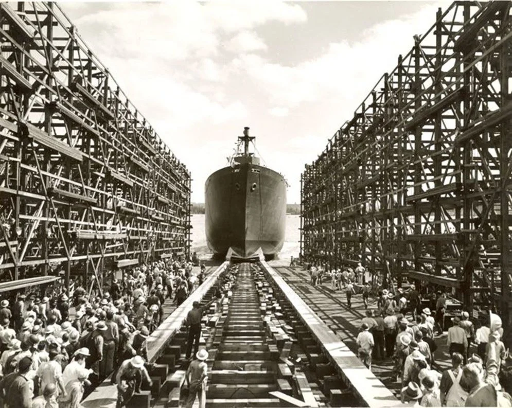 Black and white archival photo: Workers gather around large scaffold structures and tracks to launch a WWII Liberty Ship.